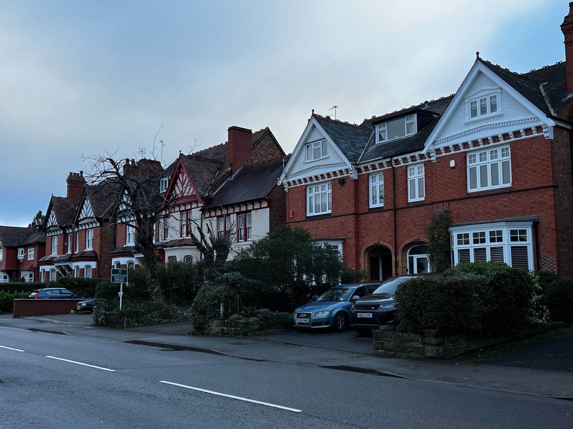 Row of brick houses with gabled roofs along a street; cars parked in front. Overcast sky.