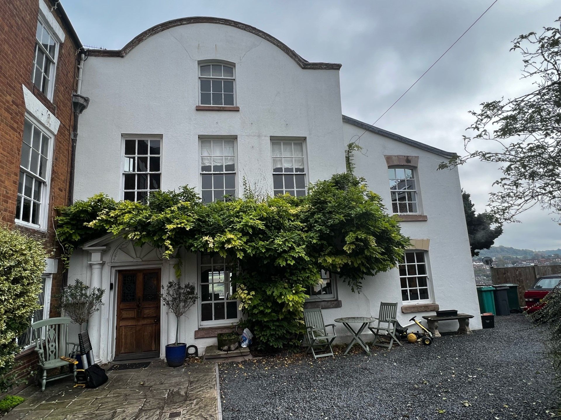 White house with arched facade, overgrown with green vines, set on a gravel driveway.
