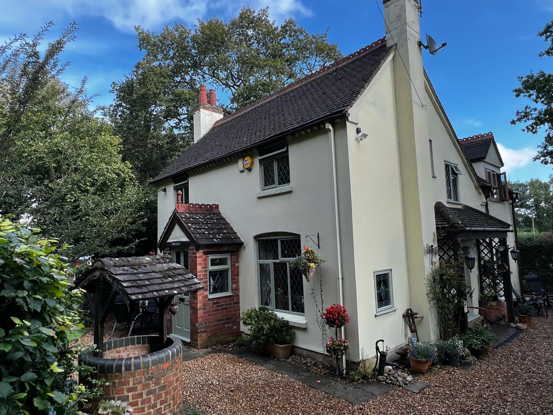 Cottage with light-colored walls, a dark roof, and a brick well in front. Blue sky and trees surround the house.