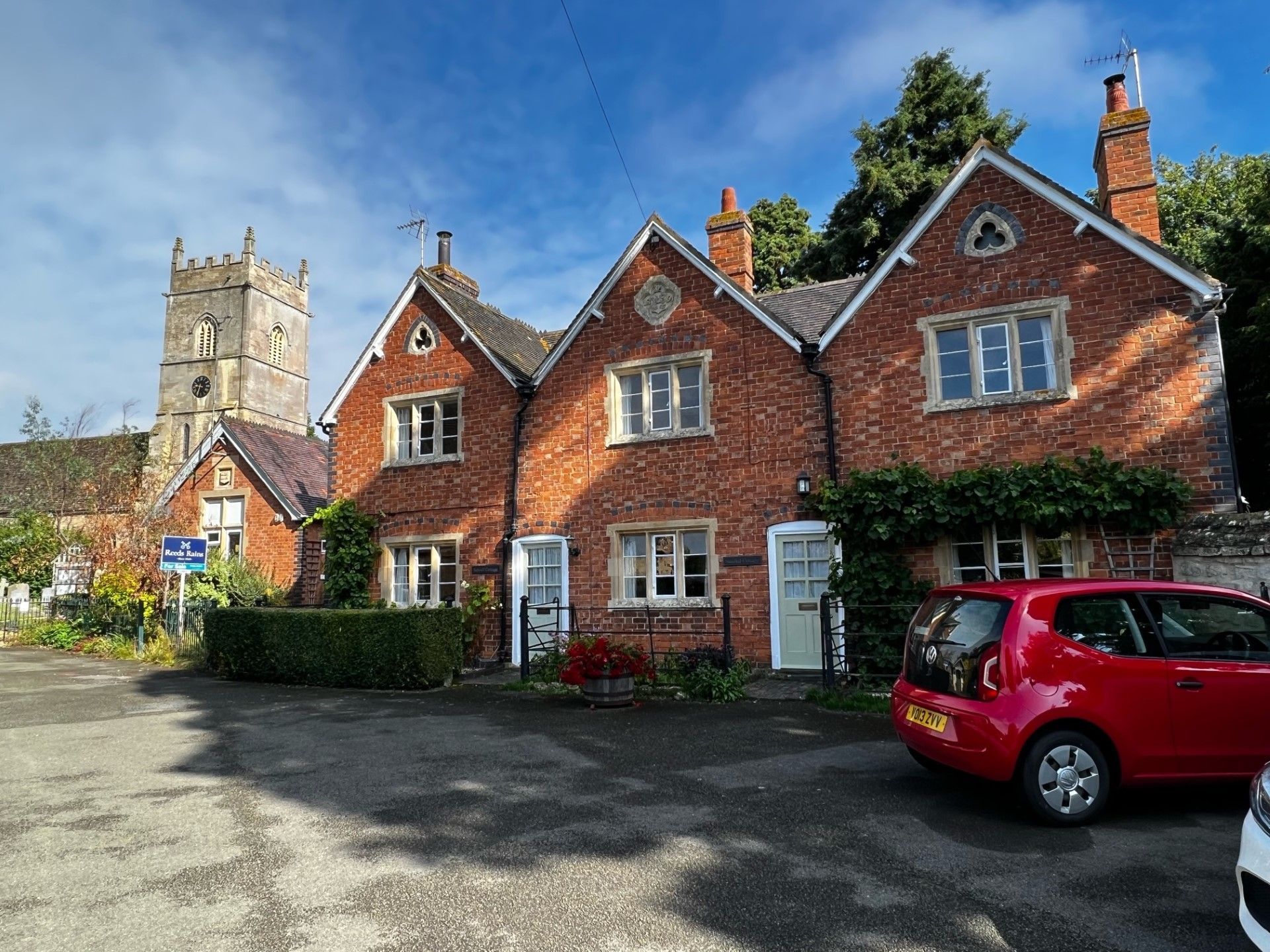 Red brick cottages with a church tower in the background. A red car is parked nearby on a sunny day.