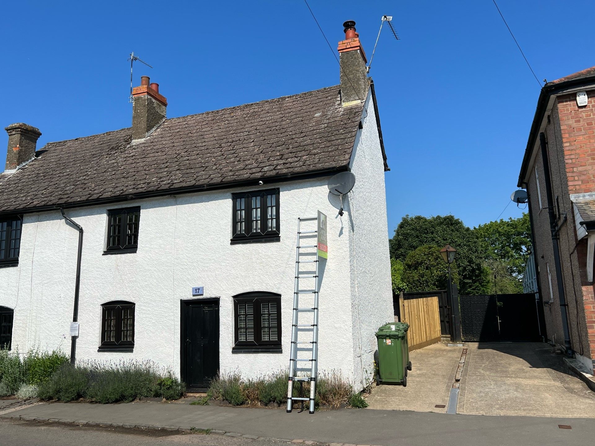 White cottage with black door and windows, ladder propped against it, green bin, driveway, and blue sky.