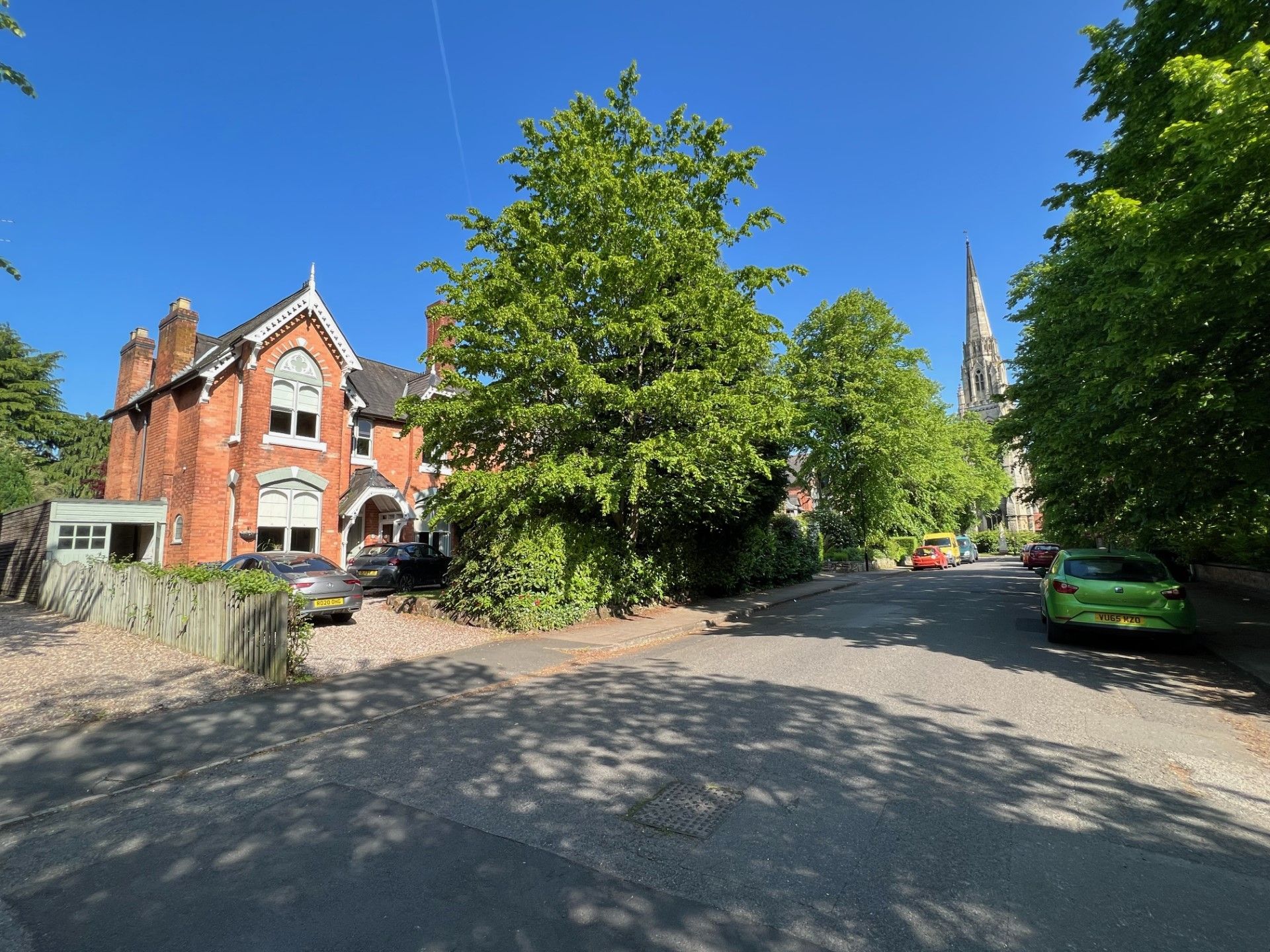 Red brick building on a street, green trees, blue sky, car parked.