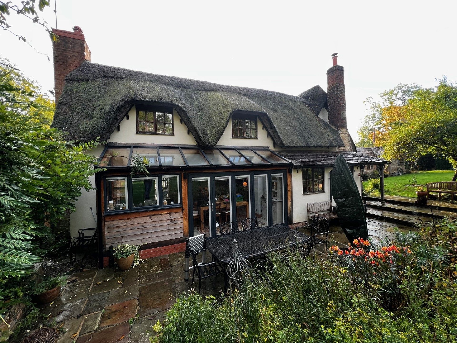 Thatched cottage with brickwork, stone patio, and surrounding foliage.