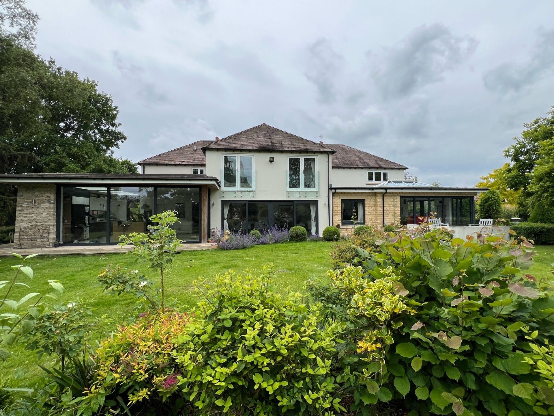 Rear view of a two-story house with glass extensions, overlooking a lush green garden under a cloudy sky.