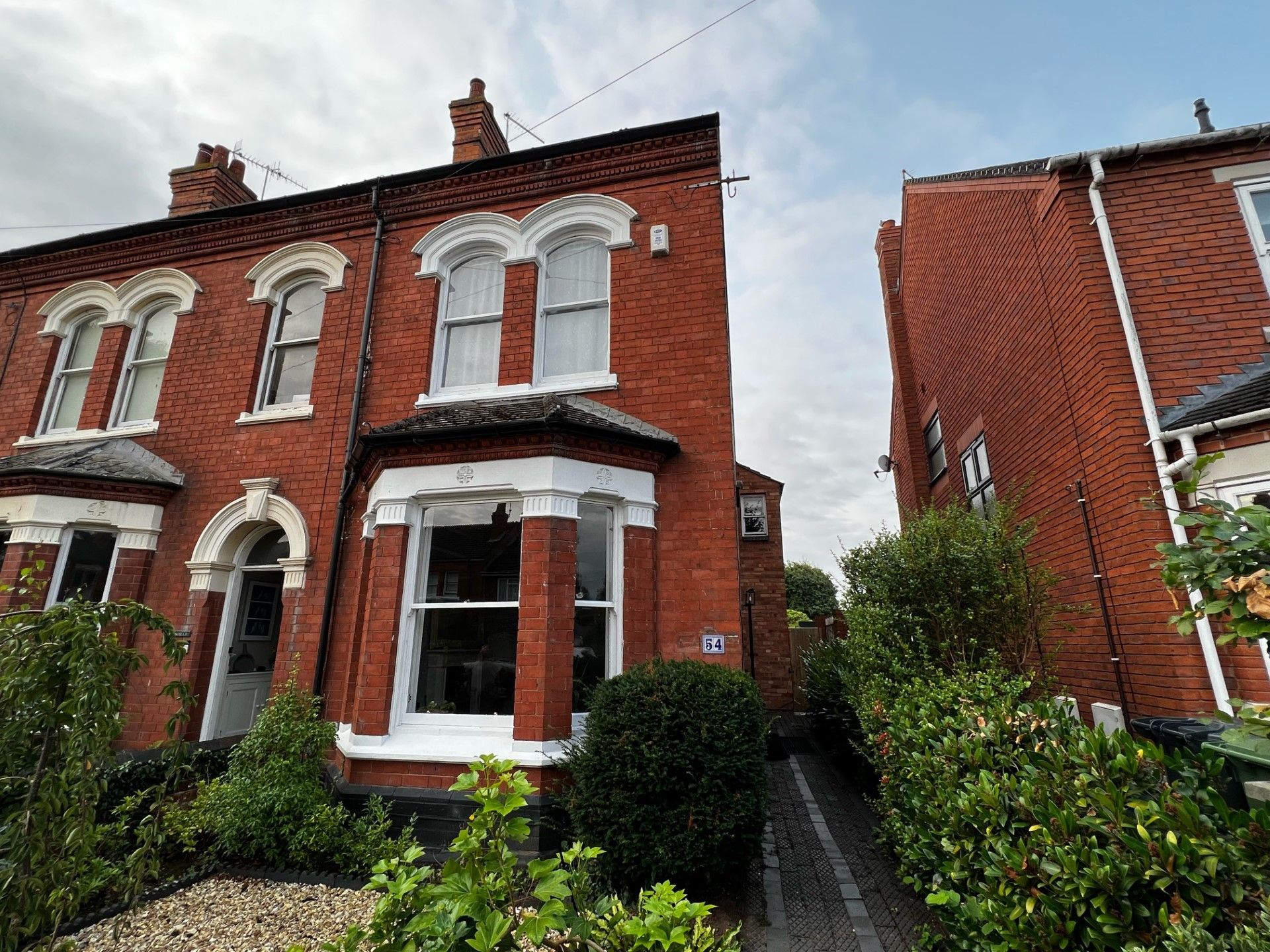 Red brick terraced houses with white window frames and bushes in front under a cloudy sky.