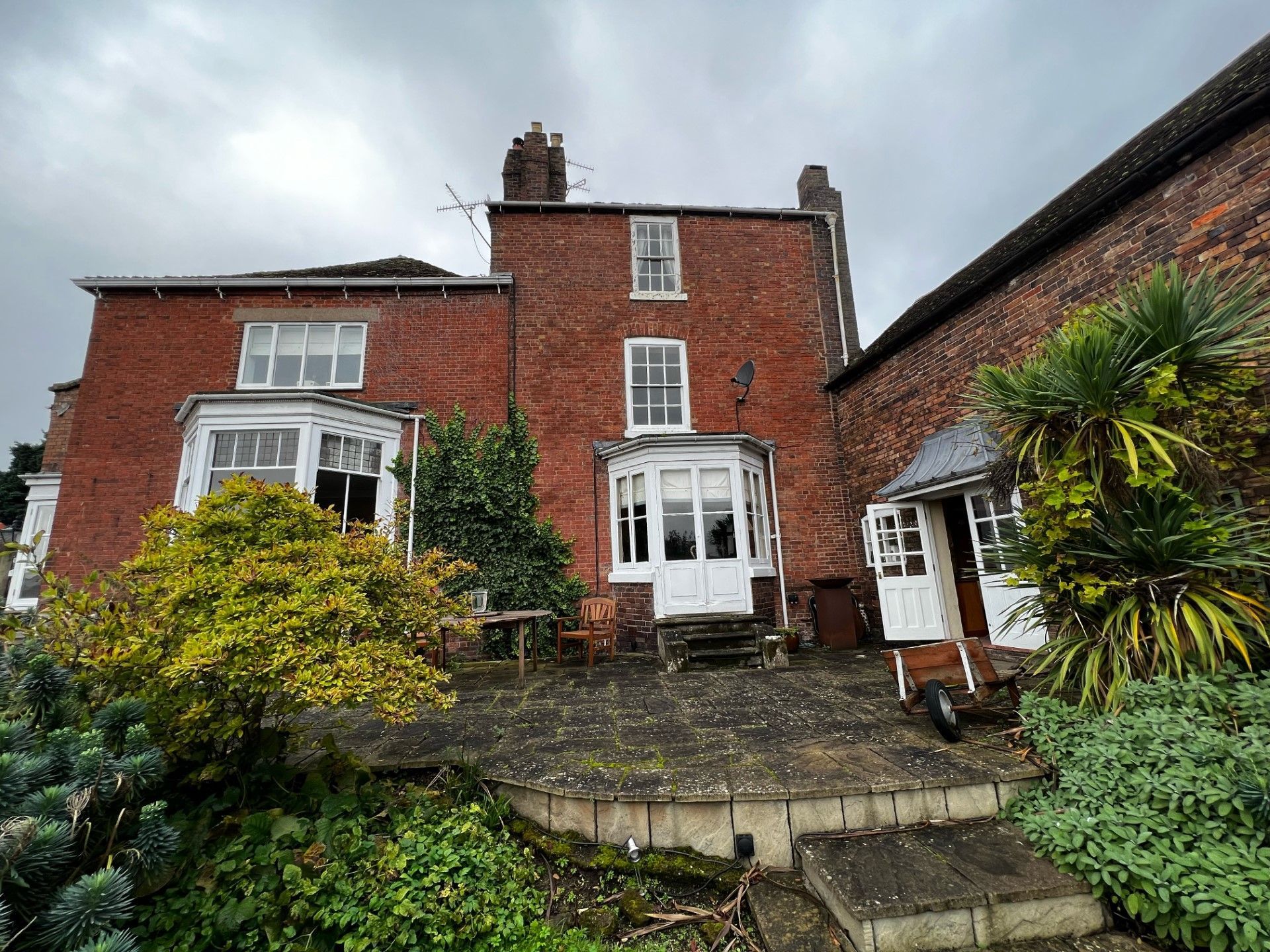 Red brick house with white trim, overgrown garden, cloudy sky.