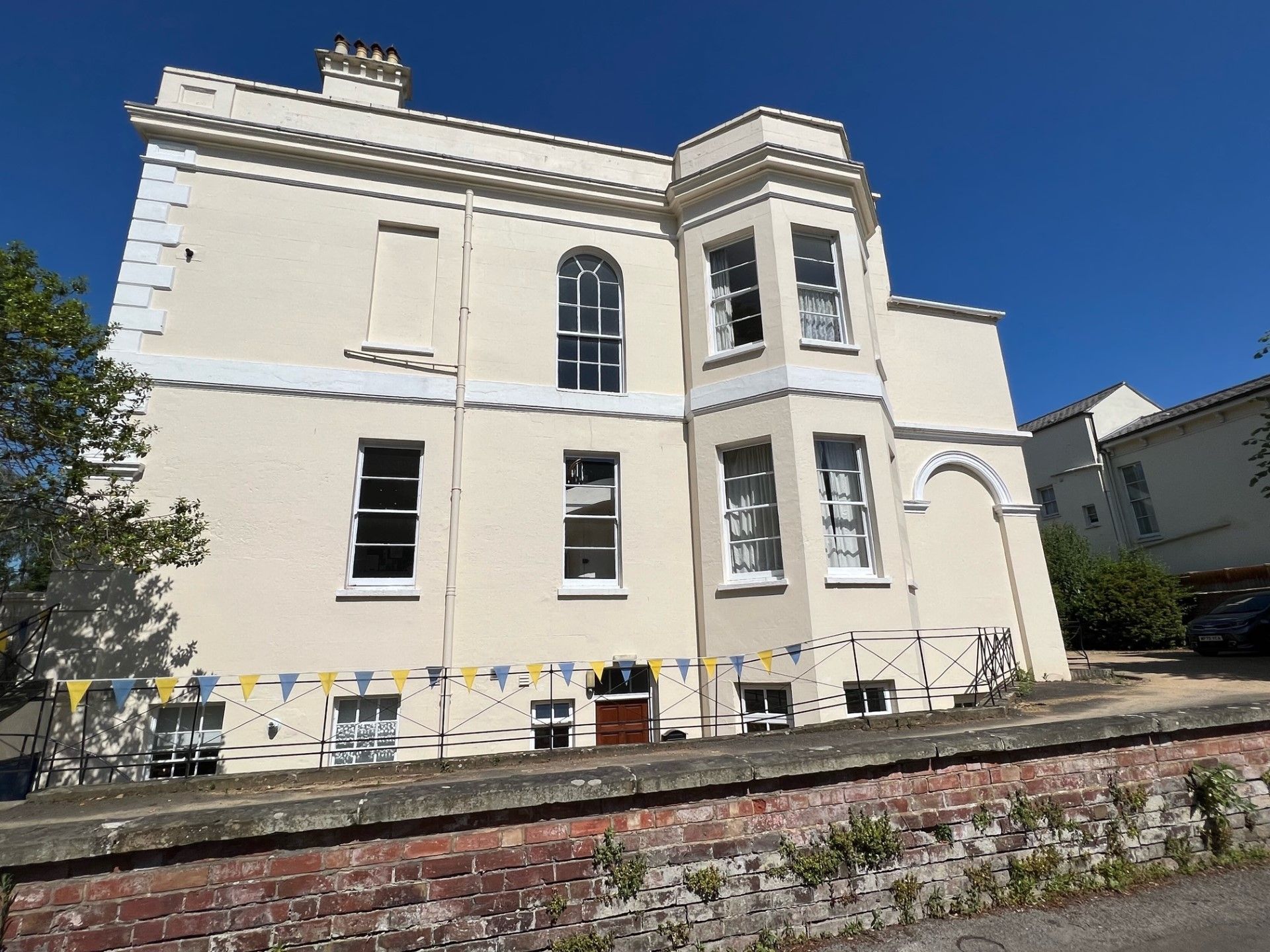 Cream-colored building with a bay window, surrounded by a blue sky, brick wall, and bunting.