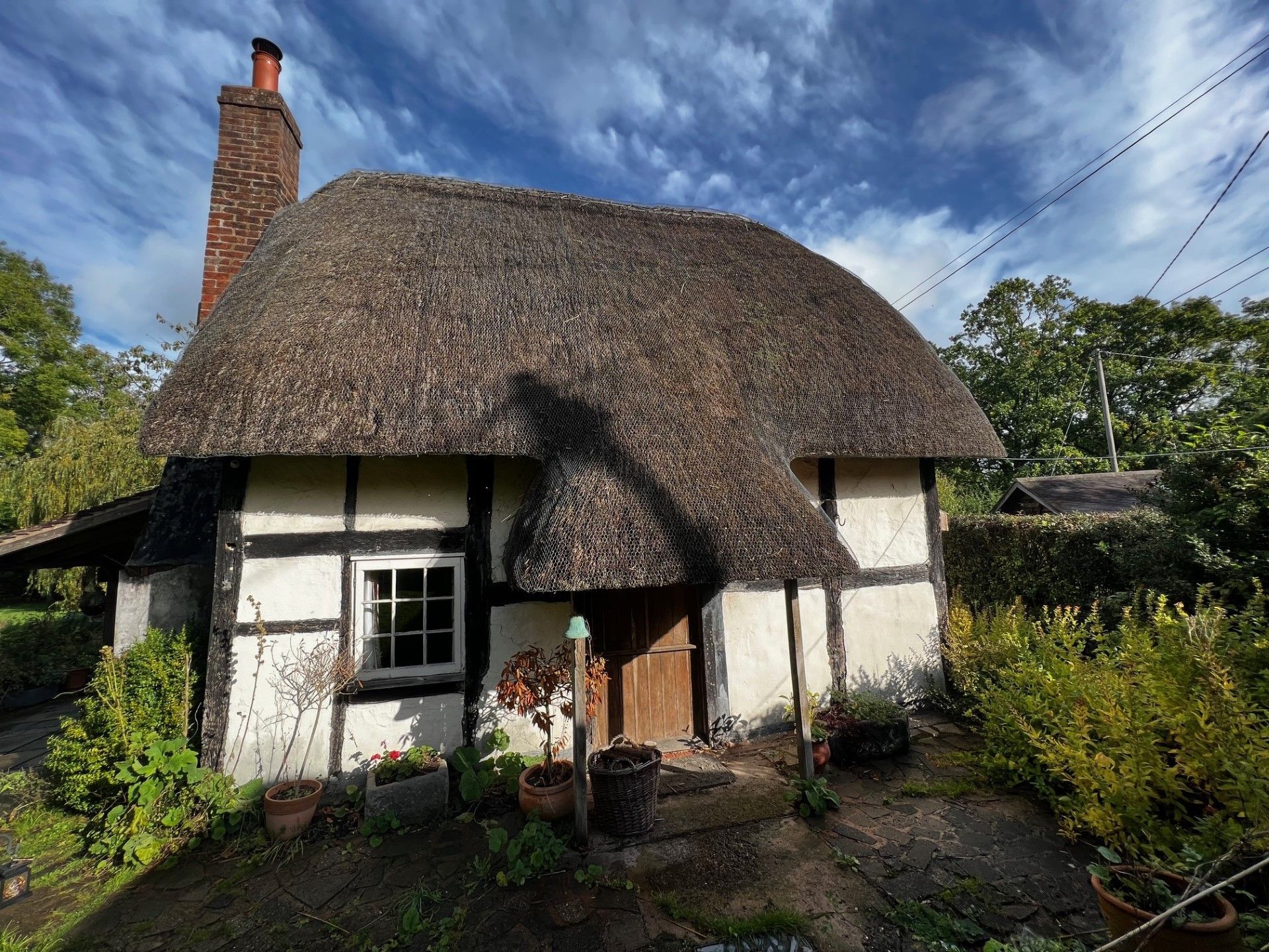 Thatched-roof cottage with white walls, dark beams, and a chimney, surrounded by greenery under a blue sky.