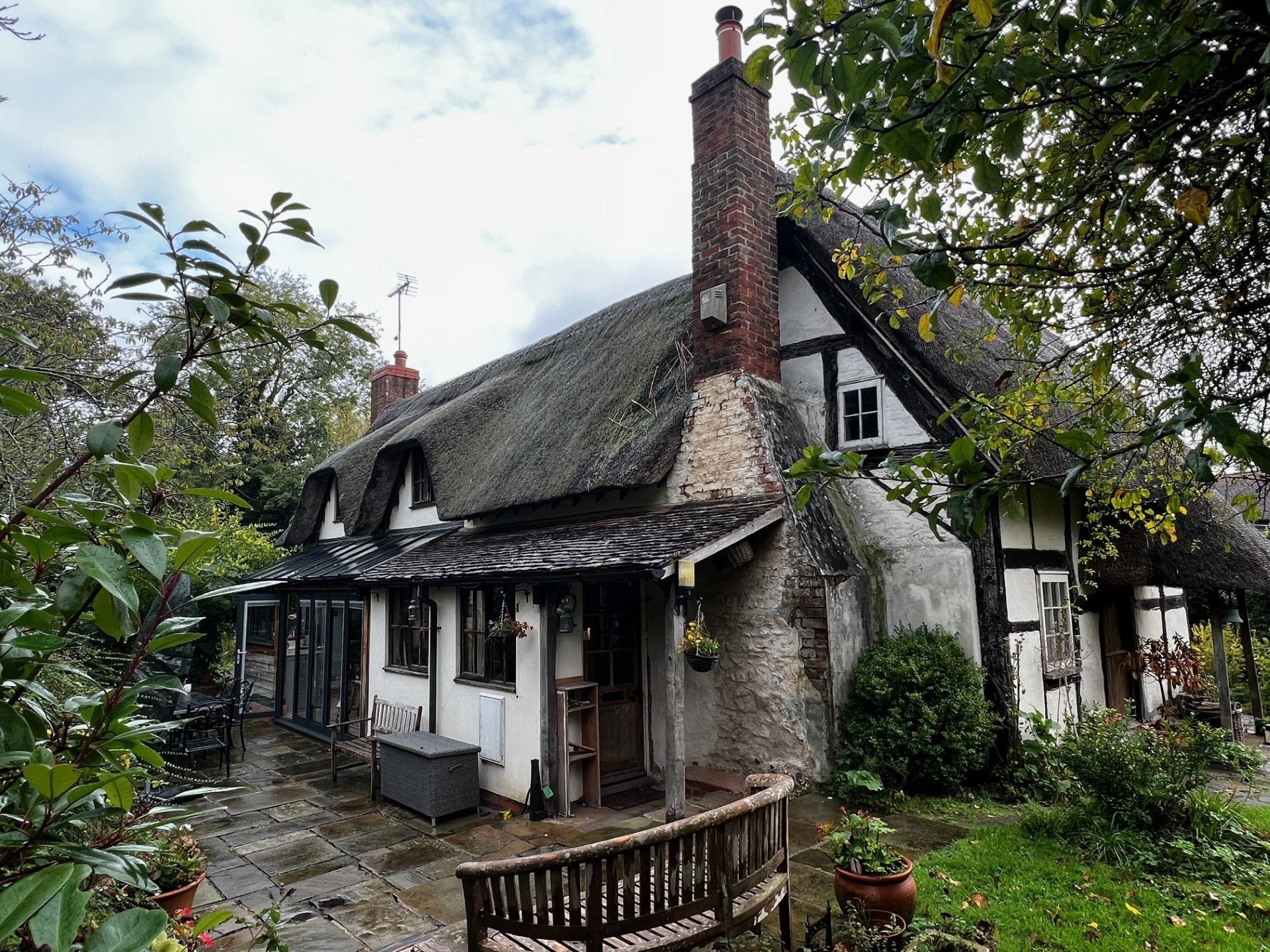 Thatched-roof cottage, white and black timber frame, brick chimney, surrounded by greenery.