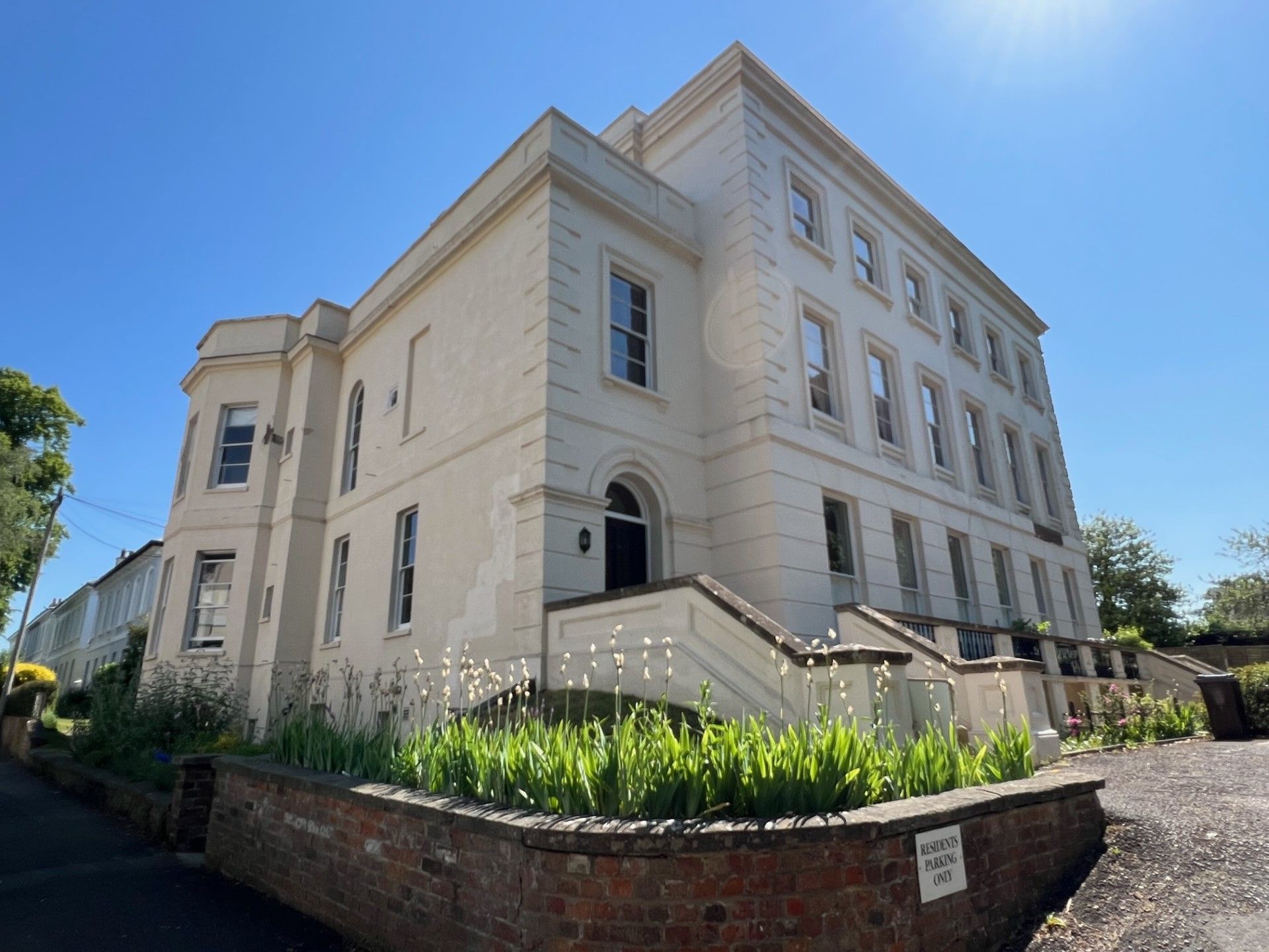 Cream-colored multi-story building with many windows, a small garden, and a brick wall on a sunny day.