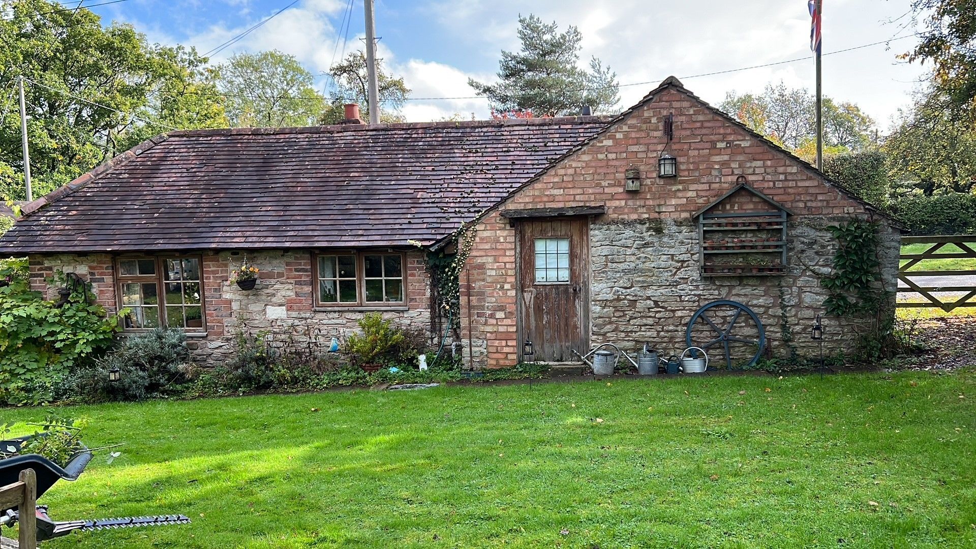 Quaint brick cottage with a grass lawn, wooden door, and boarded window; sunny outdoor setting.