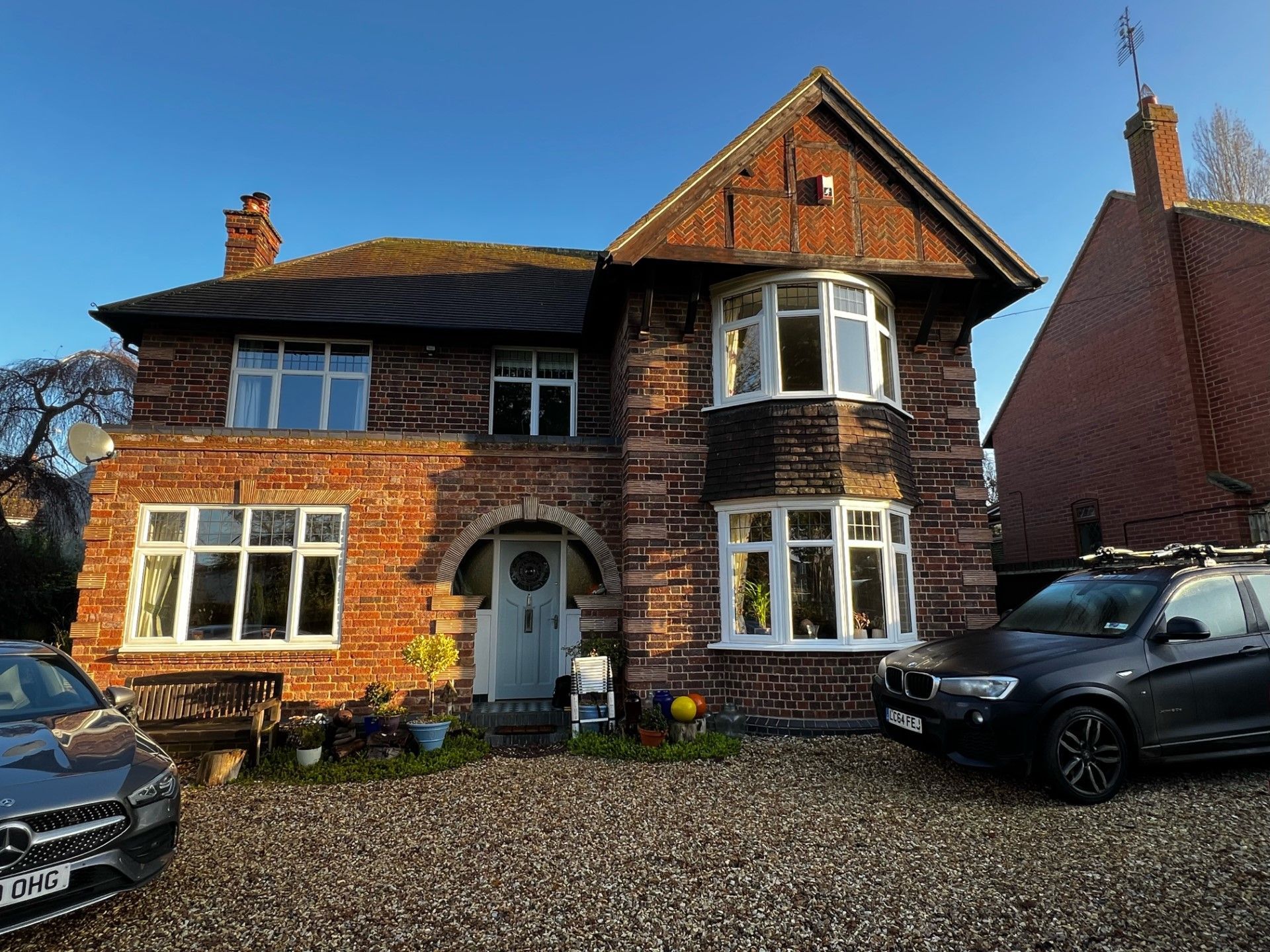 Two-story brick house with bay windows, gravel driveway, and two cars parked in front under a blue sky.