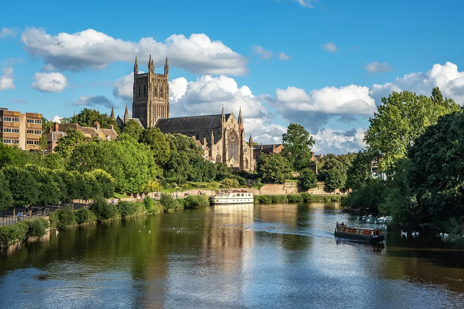 River scene with a large church, boats, trees, and buildings under a blue sky with clouds.