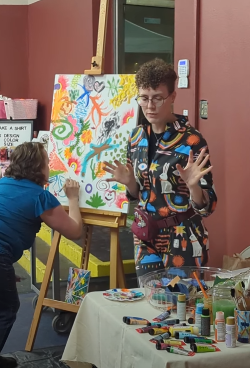 Lore in a Mx. Frizzle-esque jumpsuit guiding a community expressive art project at the Tacoma Night Market. 