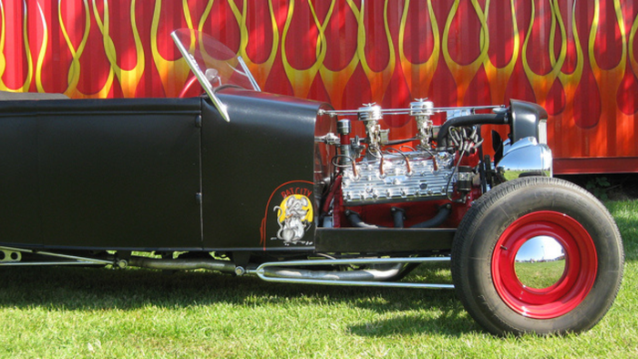 A black car with red wheels is parked in the grass in front of a trailer with flames painted on it.