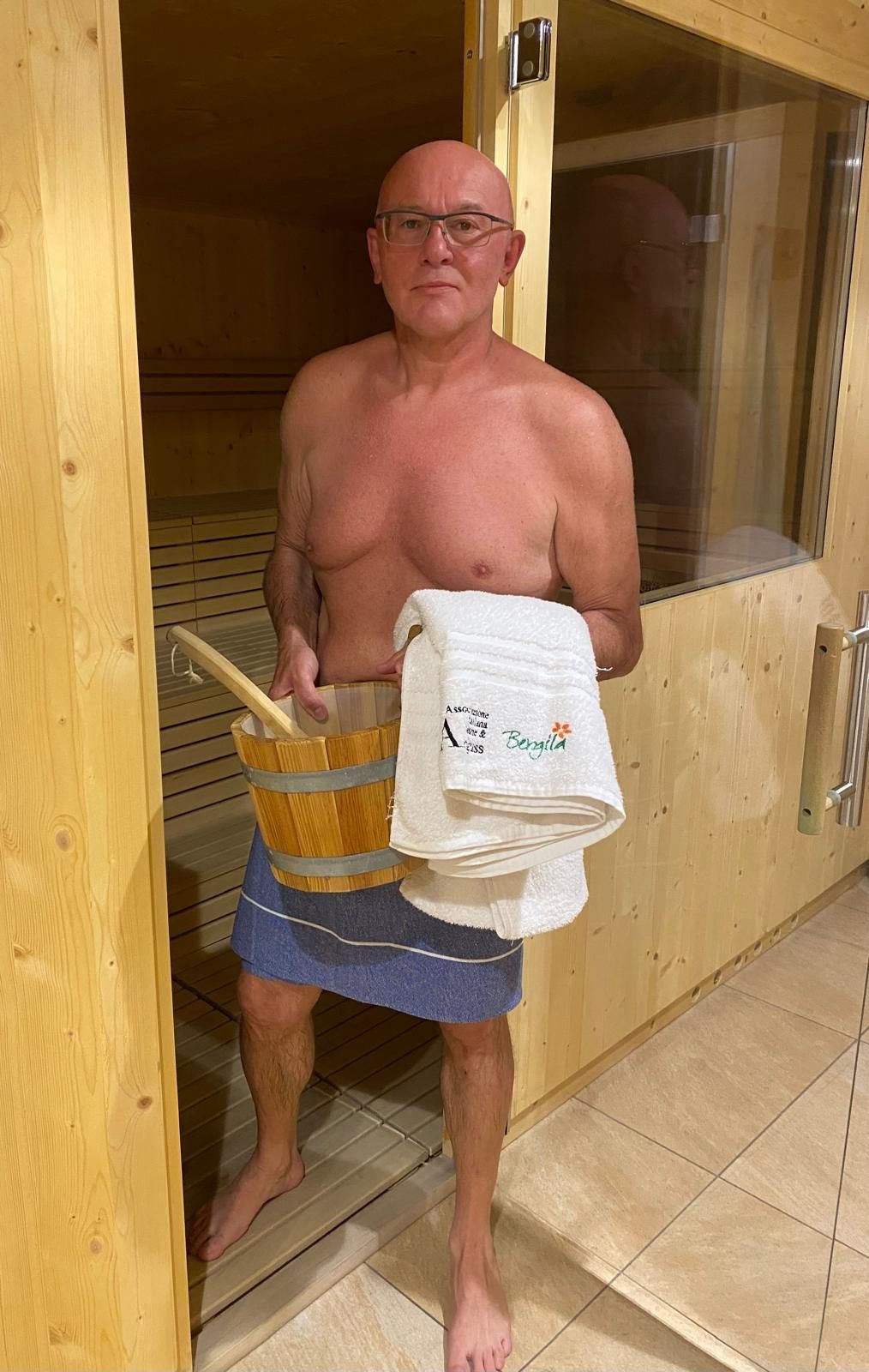 Un uomo è in piedi sulla soglia di una sauna, con in mano un asciugamano e un secchio, e alle sue spalle si vedono le pareti di legno della sauna.