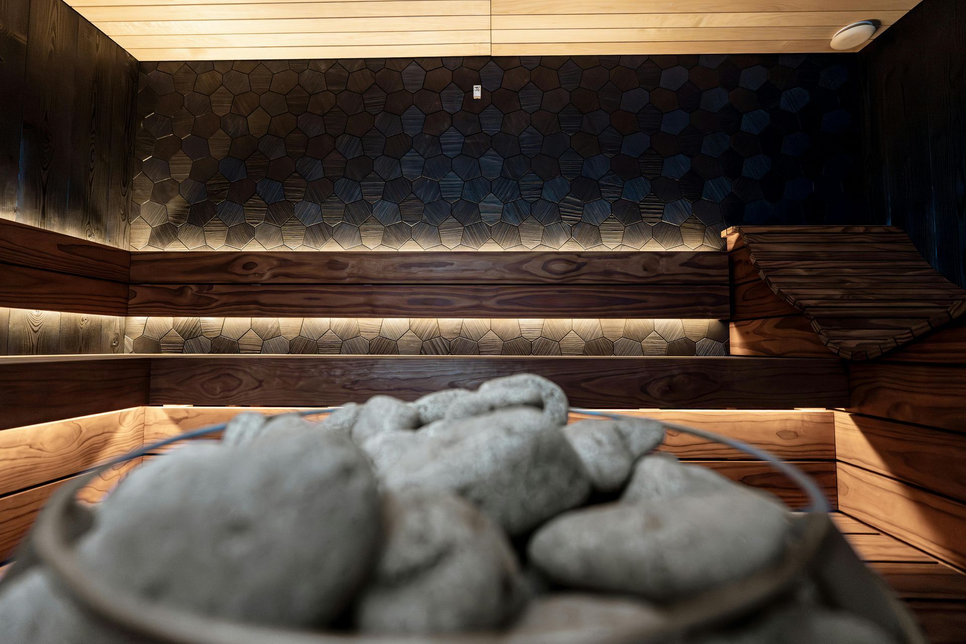 Interno di una sauna in legno con luce calda e un mucchio di pietre grigie lisce in primo piano.
