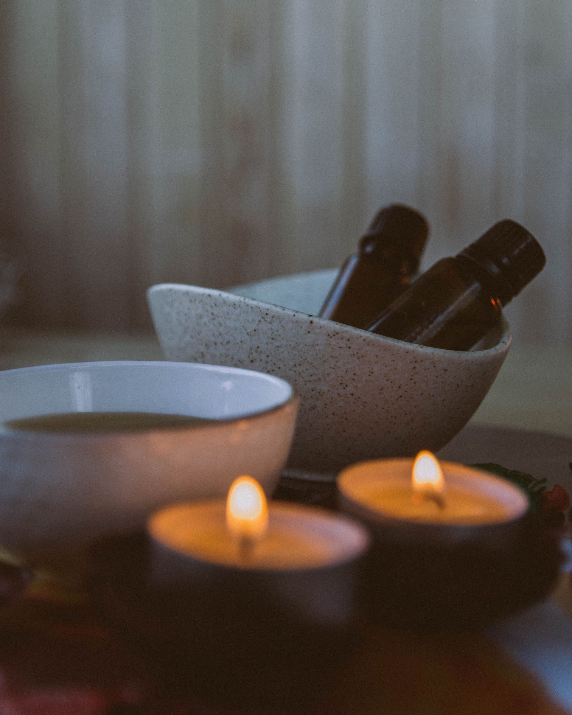 Scena da spa con candele accese, una ciotola bianca e bottiglie nere accanto a una tenda sullo sfondo.