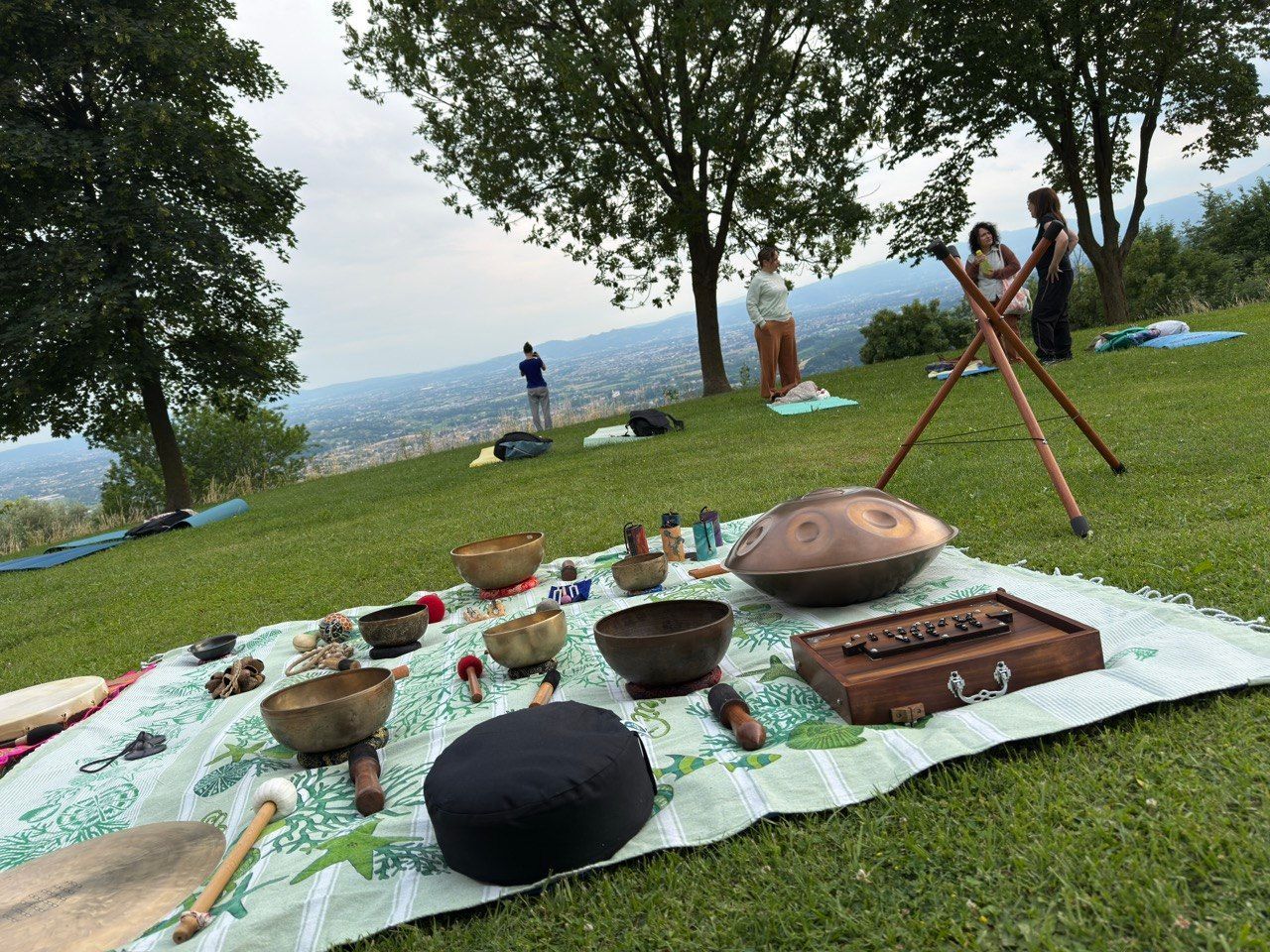 Coperta da picnic all'aperto con campane tibetane, tamburo e treppiede su una collina erbosa sotto gli alberi.