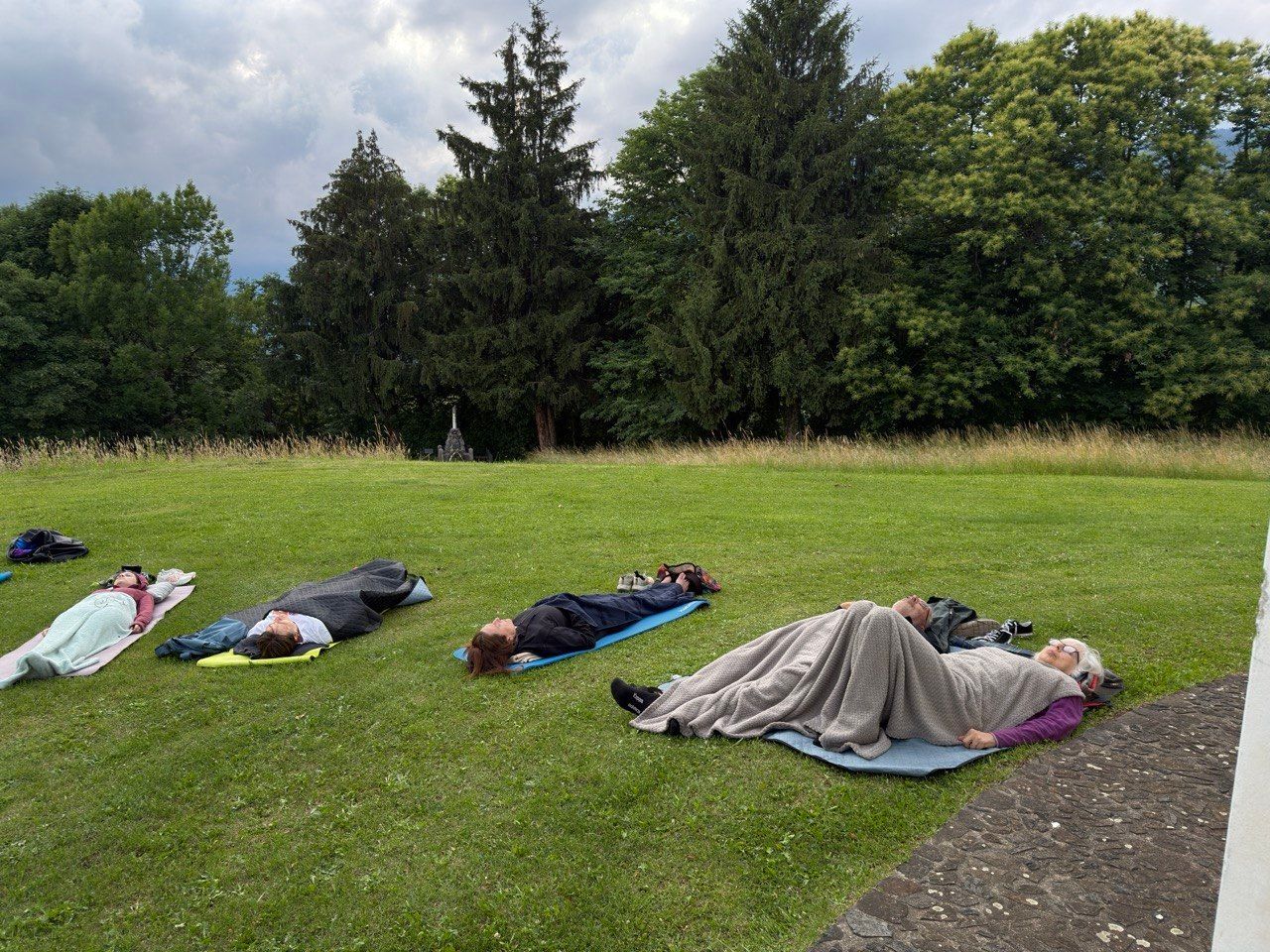 Persone sdraiate su coperte in un parco erboso, che si riposano all'ombra degli alberi.