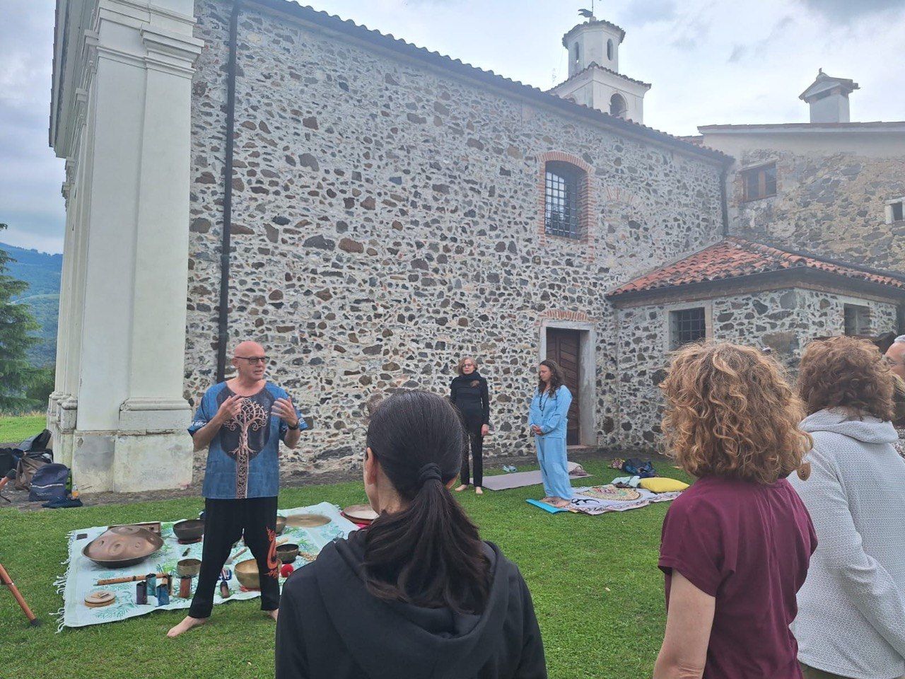 Persone che ascoltano un altoparlante all'aperto accanto a una chiesa in pietra, con un pannello informativo sull'erba.