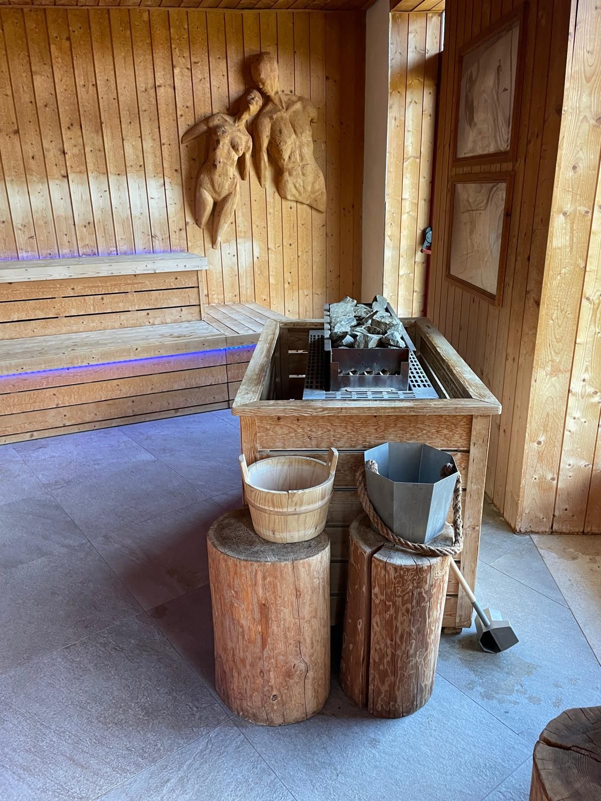 Stanza sauna in legno con panca, secchio, mestolo e un tavolino con piante e pietre.