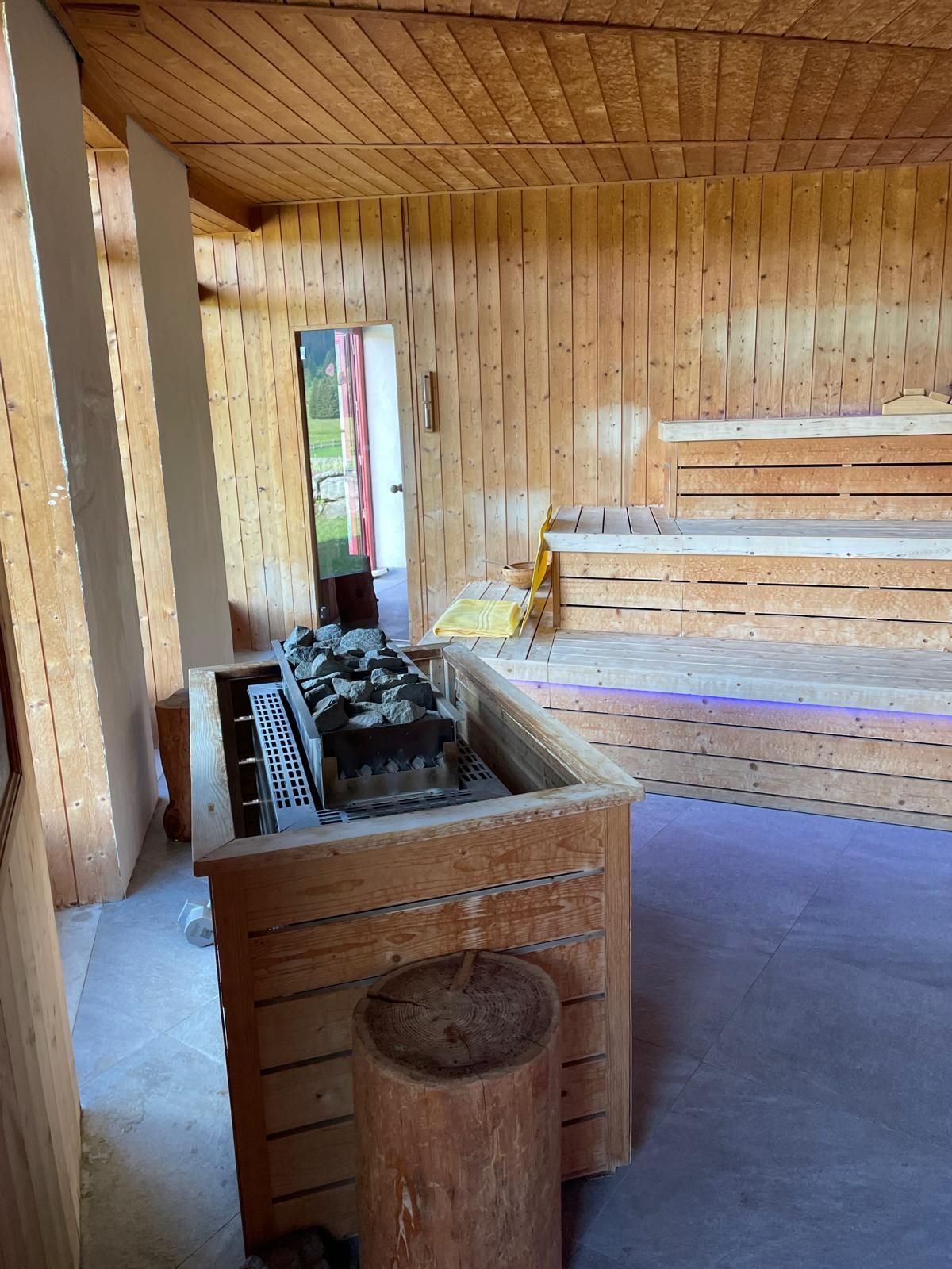 Interno di sauna in legno con panche, stufa e porta verso l'esterno.