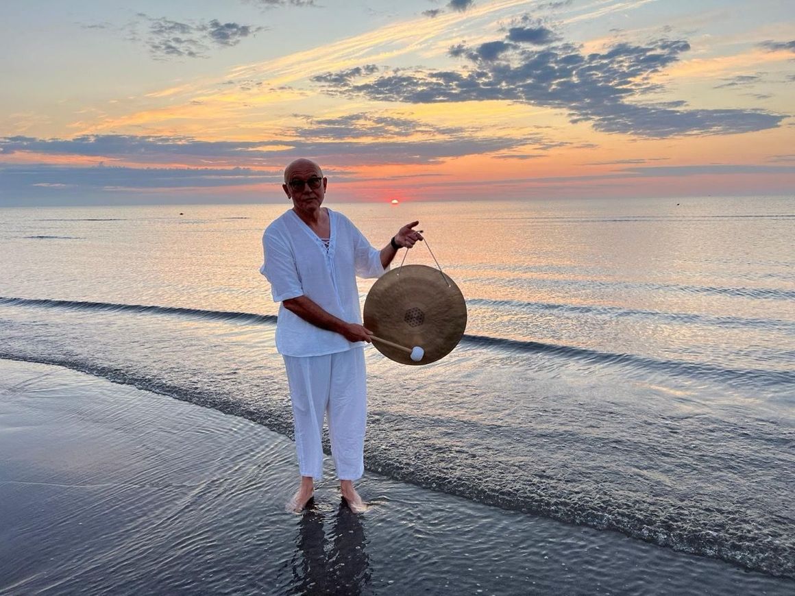 Una persona vestita di bianco, in piedi a piedi nudi sulla riva del mare, tiene in mano un gong al tramonto.
