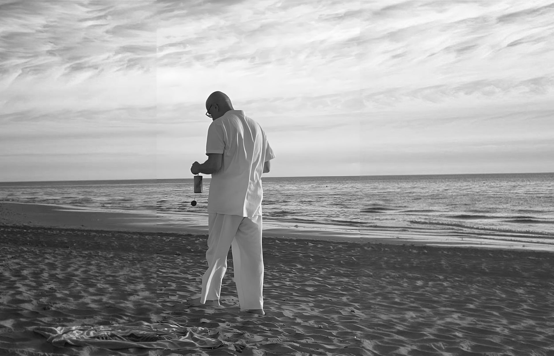 Persona vestita di bianco che cammina su una spiaggia al tramonto, con in mano una macchina fotografica, sullo sfondo le onde dell'oceano.
