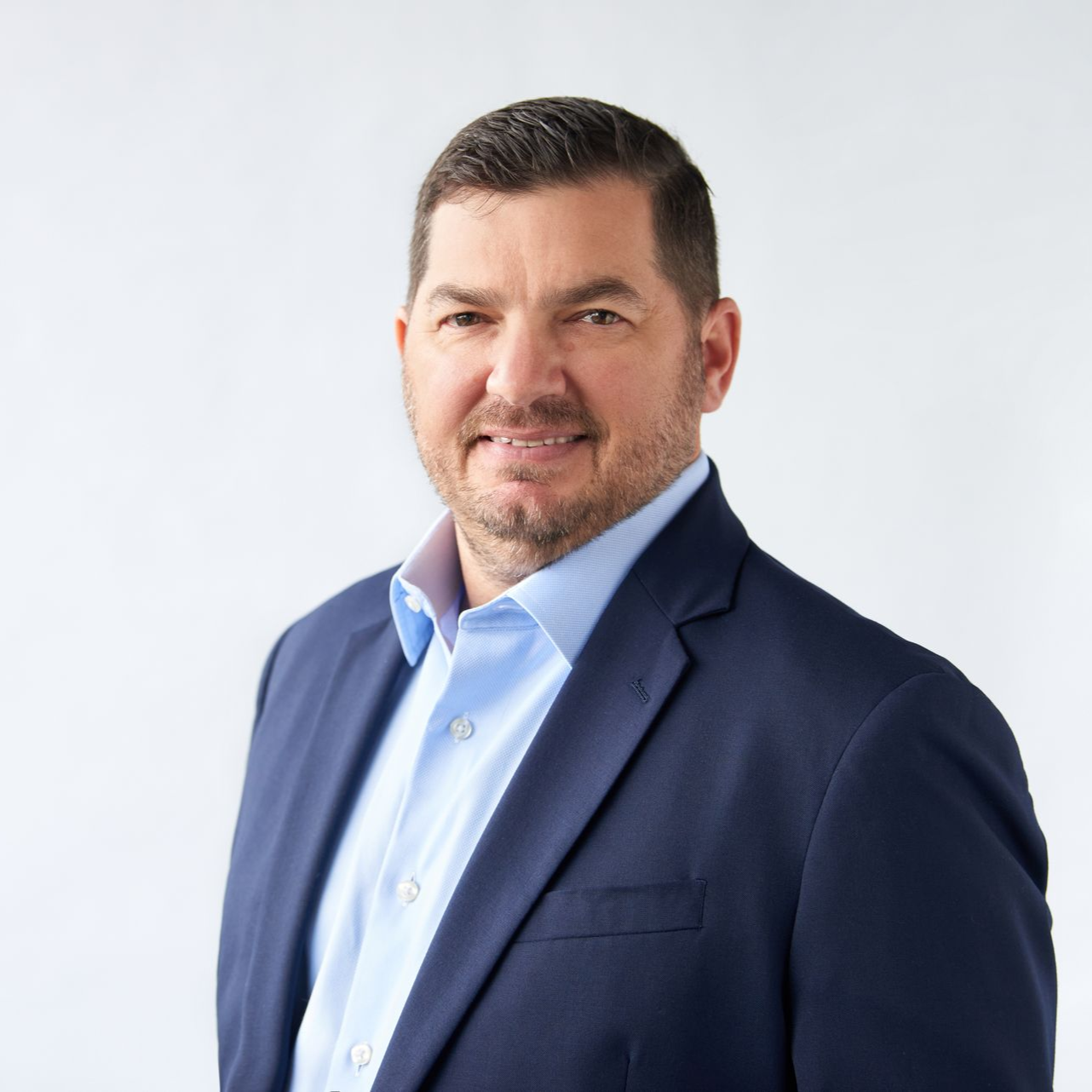 Man in a blue suit and shirt smiles at the camera. White background.