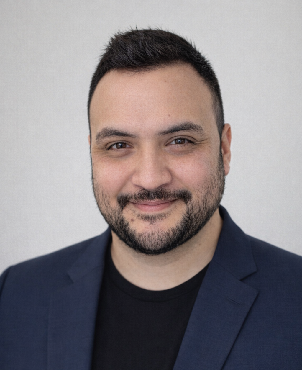Man in a dark blue jacket smiles outdoors; sunglasses hang from collar; people and wreaths in background.