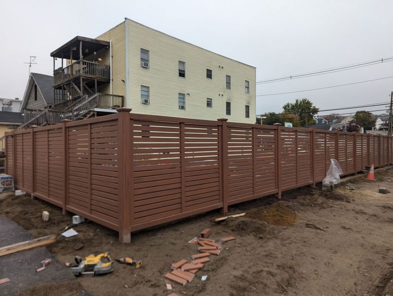 A newly installed brown horizontal-slat privacy fence stands in a dirt lot in front of a yellow three-story building.