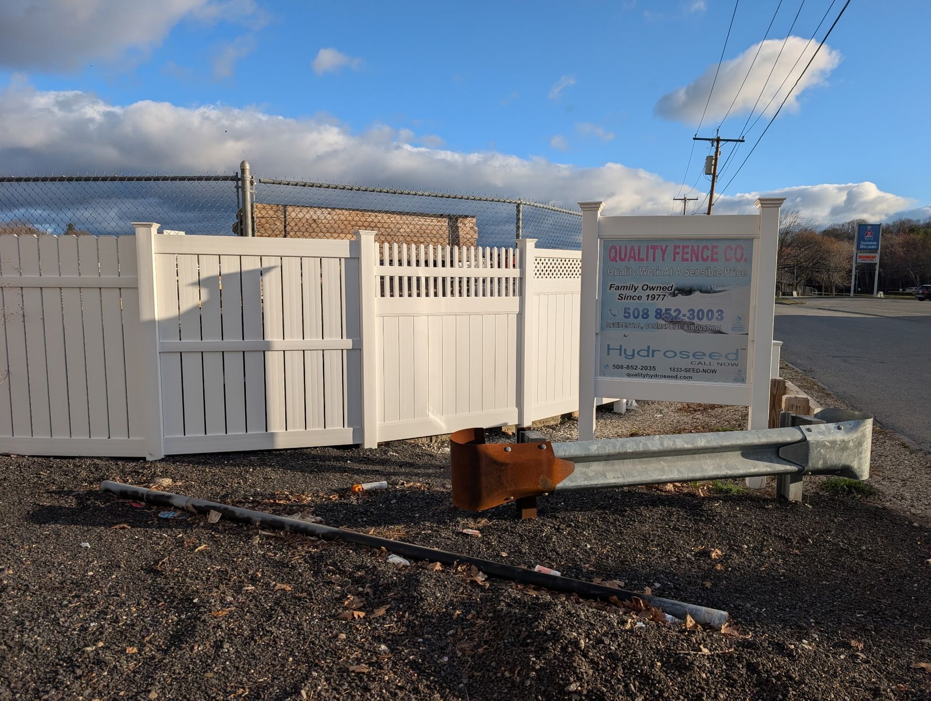 A white vinyl fence stands next to a business sign in a gravel lot under a blue sky with clouds.