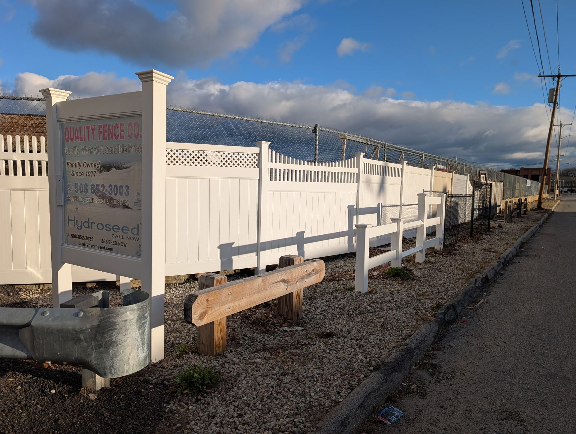 A white fence and wooden sign border a gravel lot under a cloudy blue sky, with a utility pole nearby.