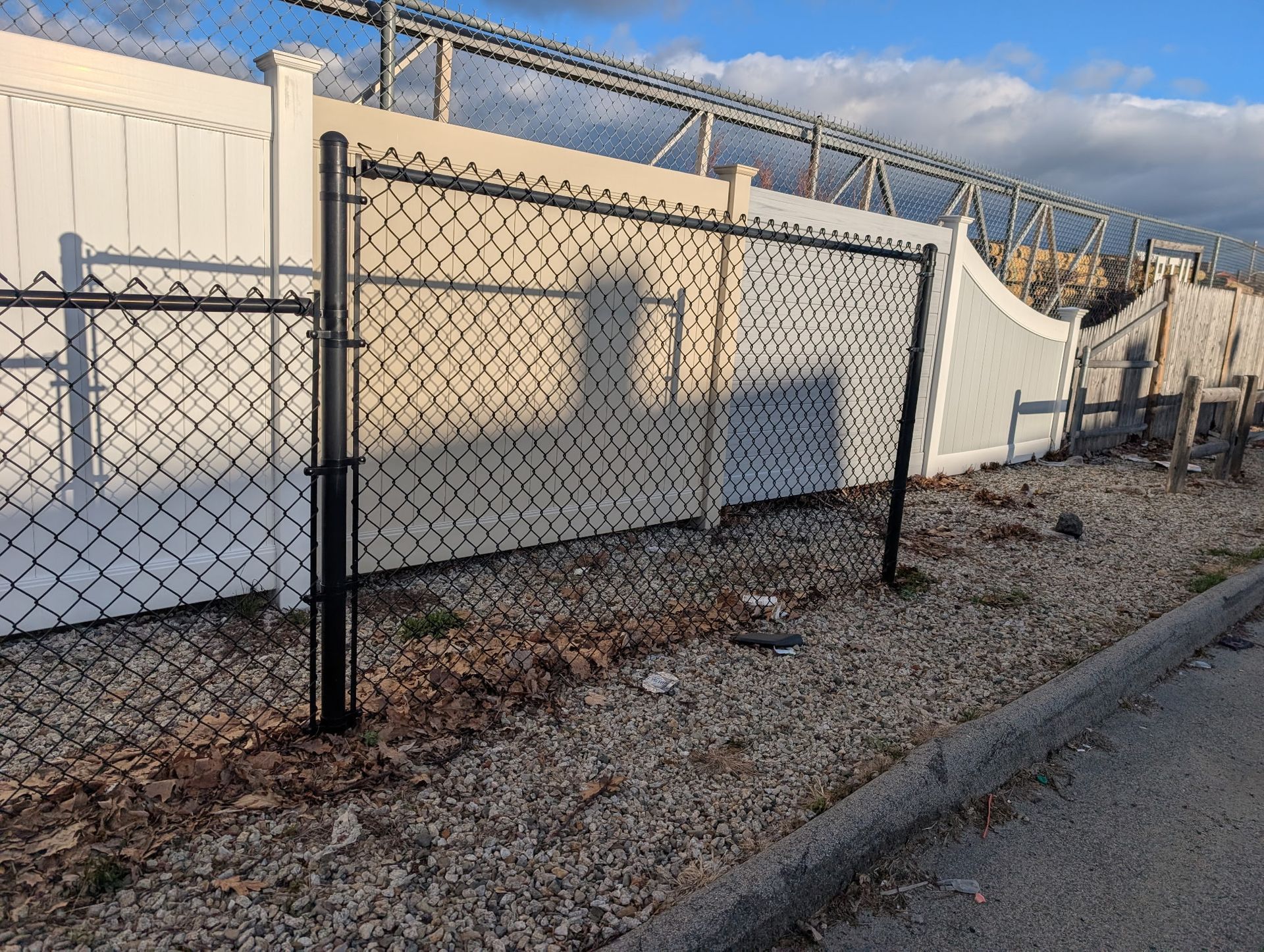 A black chain-link fence installed in front of a white vinyl fence on a gravel surface.