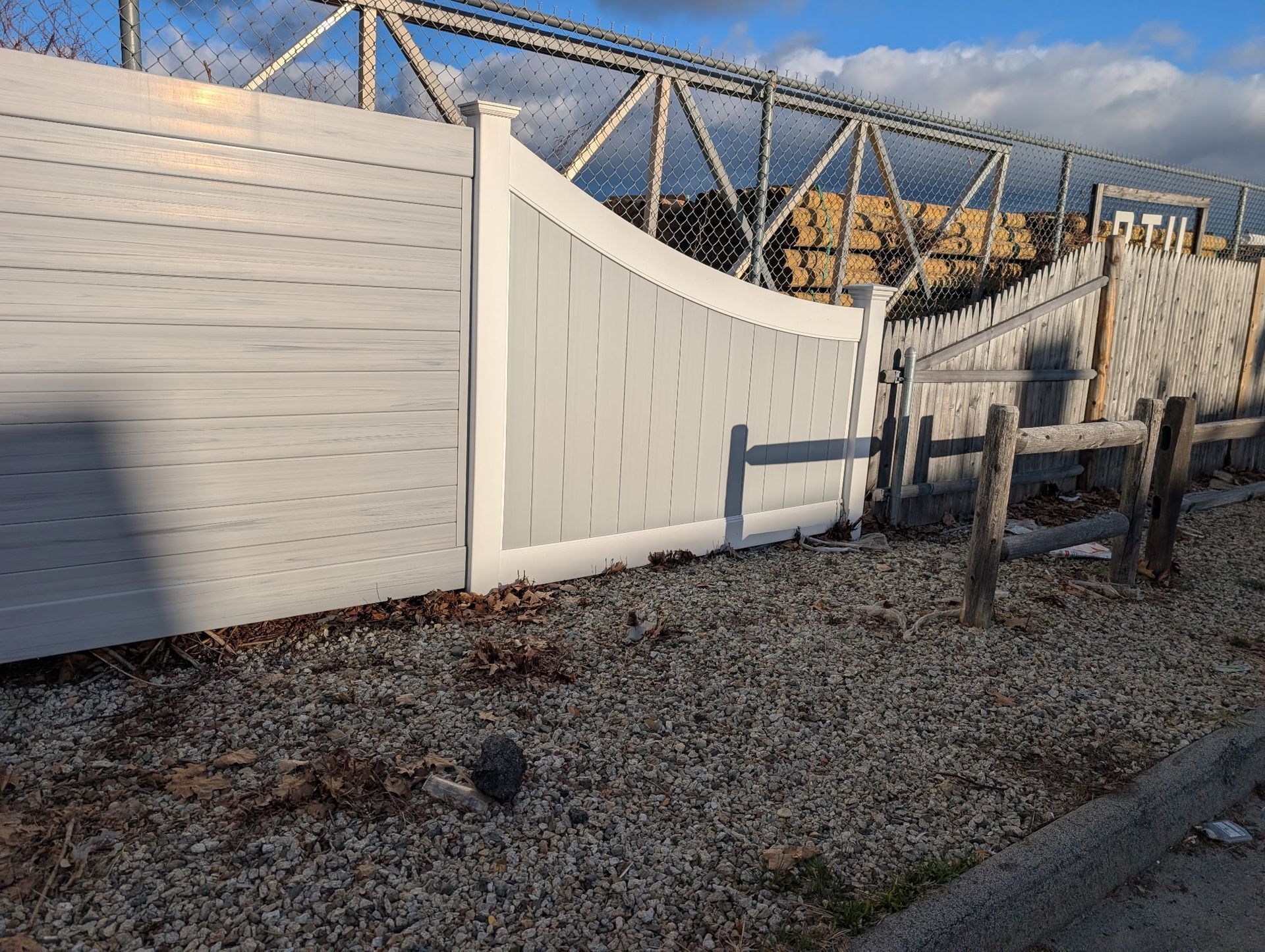 A white vinyl privacy fence with a transition panel stands above a gravel ground next to a metal chain-link security fence.