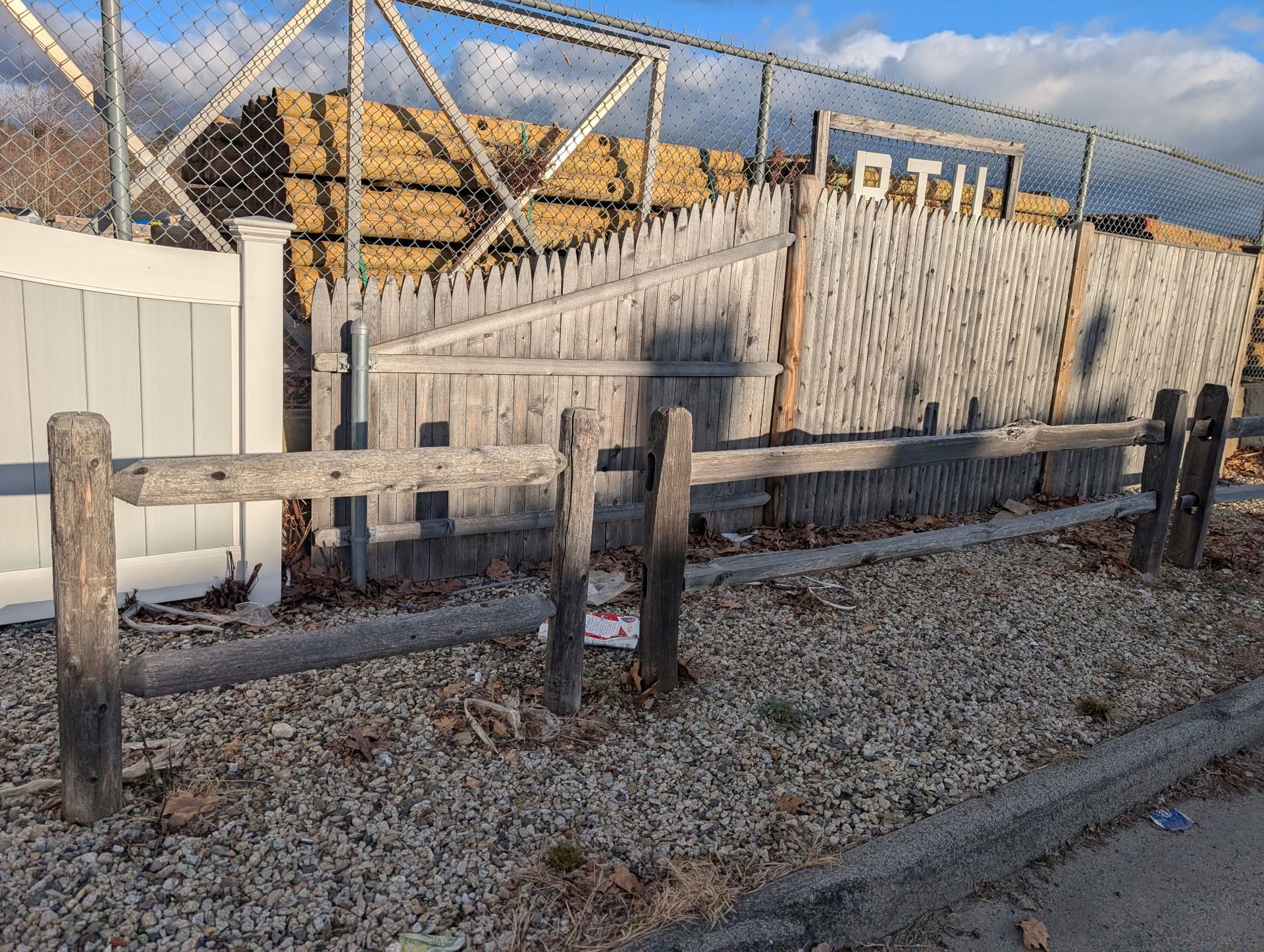A gravel foreground with a wooden post-and-rail fence in front of a chain-link fence, weathered wood fencing, and stacked logs.