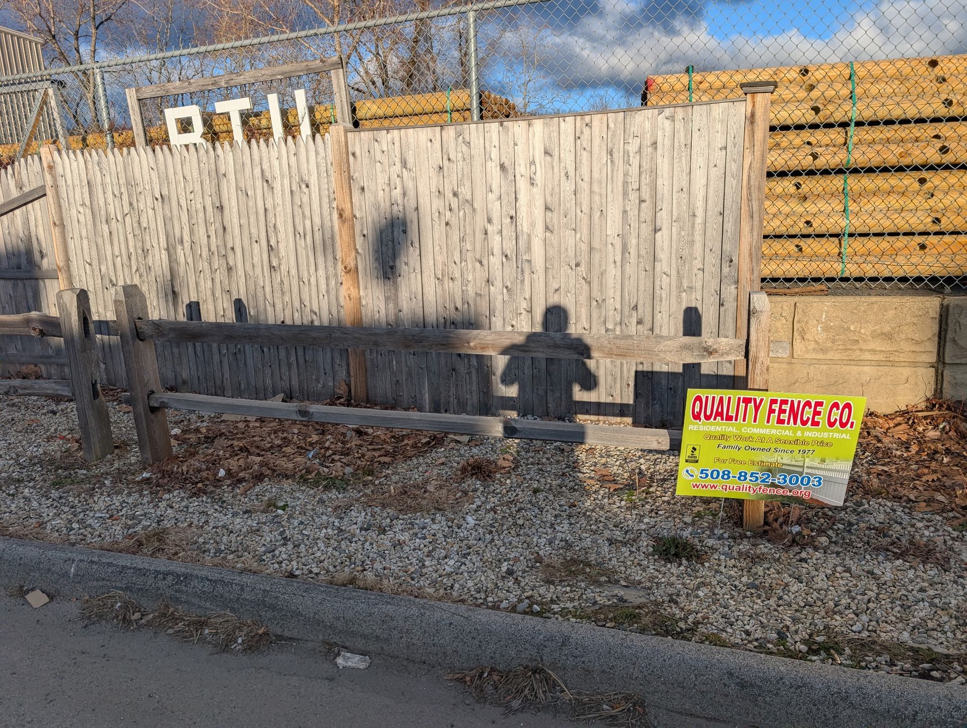 A wooden fence with a business sign for Quality Fence Co. sits on a gravel lot in front of a chain-link fence.