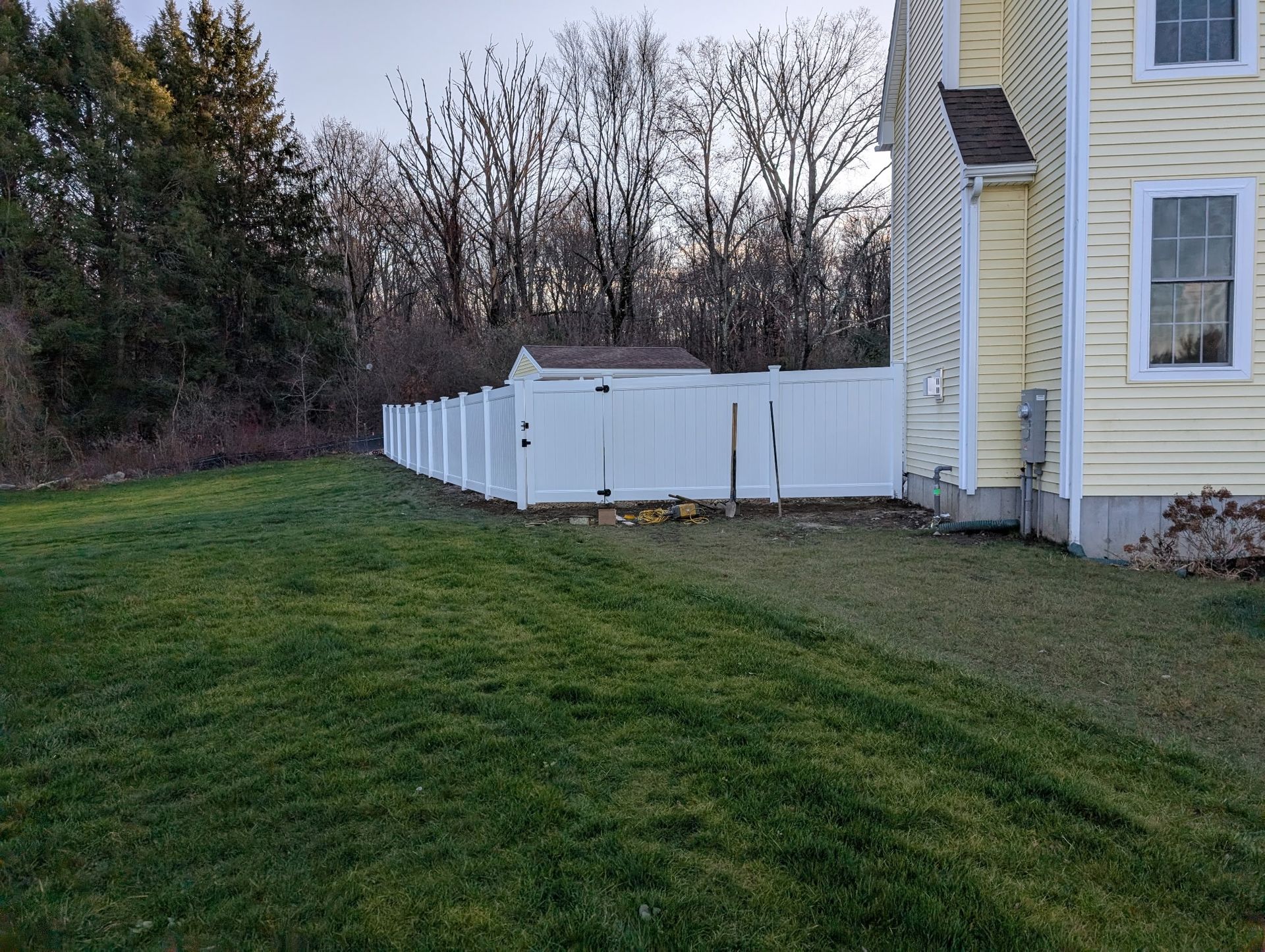 A white vinyl fence borders a grassy backyard next to a yellow house, with a small shed visible behind the fence.