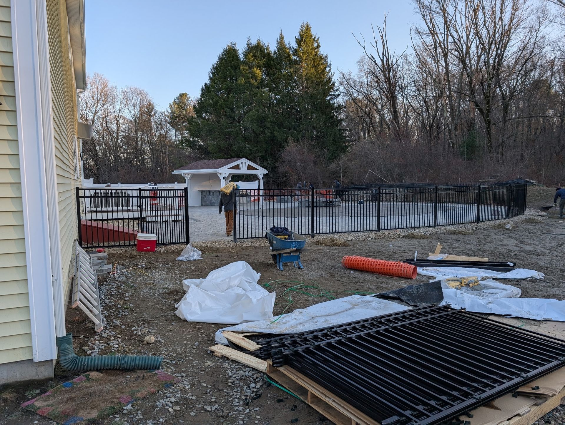 A construction site showing a house exterior, a fenced swimming pool area, and pallets of black metal fencing materials.
