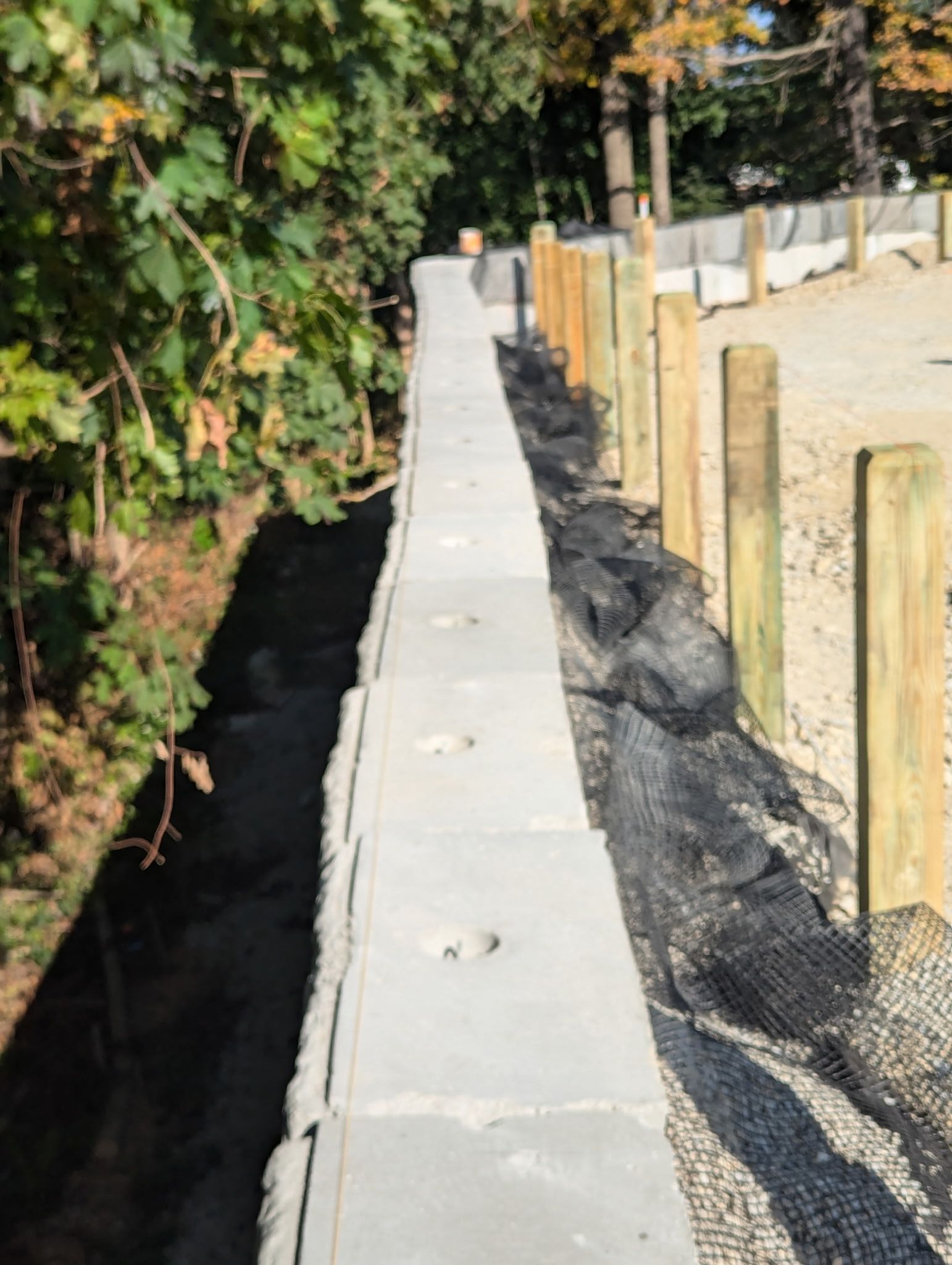 A low, modular concrete retaining wall sits next to a line of wooden fence posts and black landscape netting.