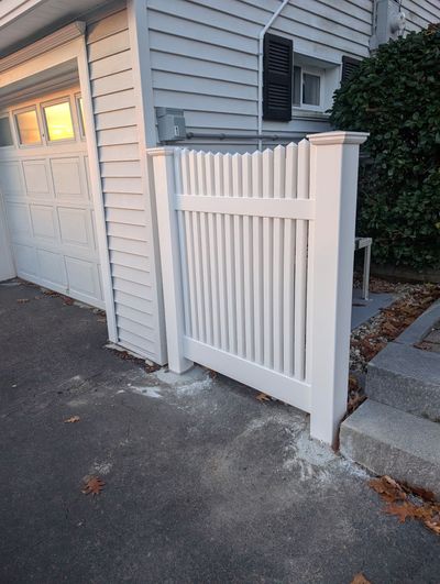 A white vinyl picket fence section is installed against the side of a house next to a garage and concrete stairs.