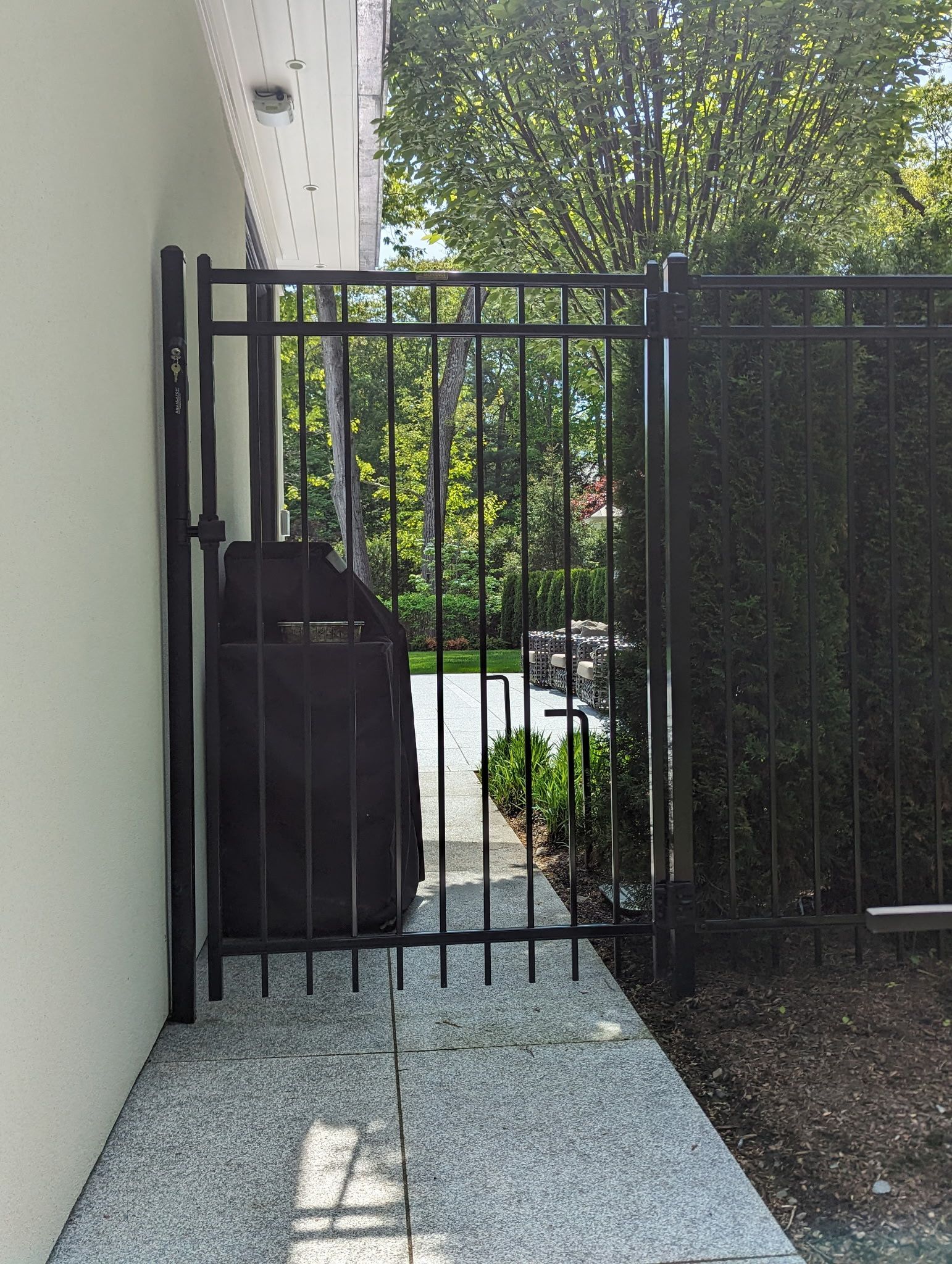 A black metal gate stands between a light-colored exterior wall and a tall hedge, leading to a paved walkway outdoors.