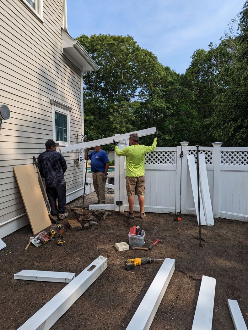 Three workers in casual clothes install a white vinyl fence in a dirt backyard next to a house with beige siding.