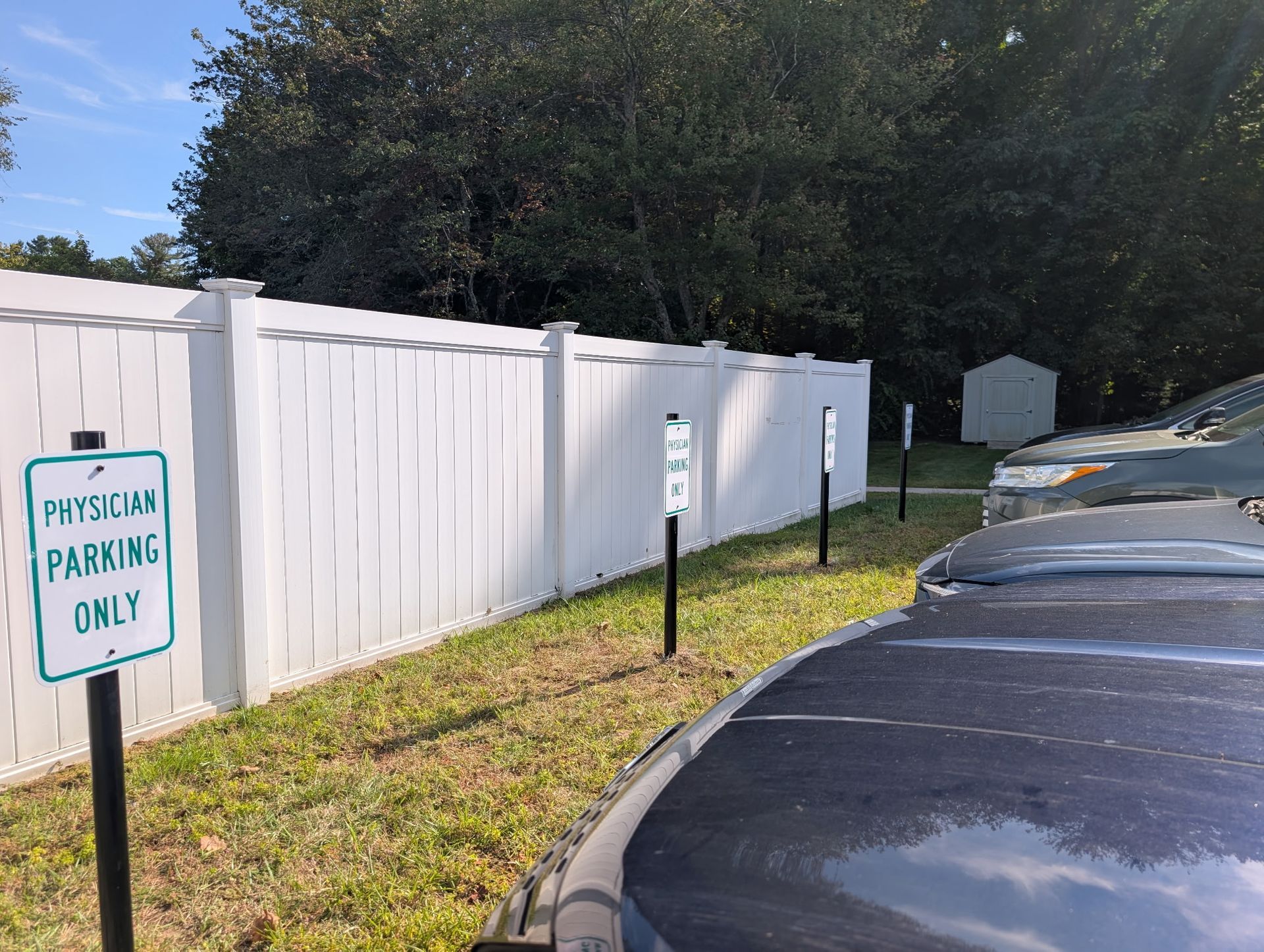 A line of Physician Parking Only signs positioned in front of a white privacy fence, with cars parked nearby.