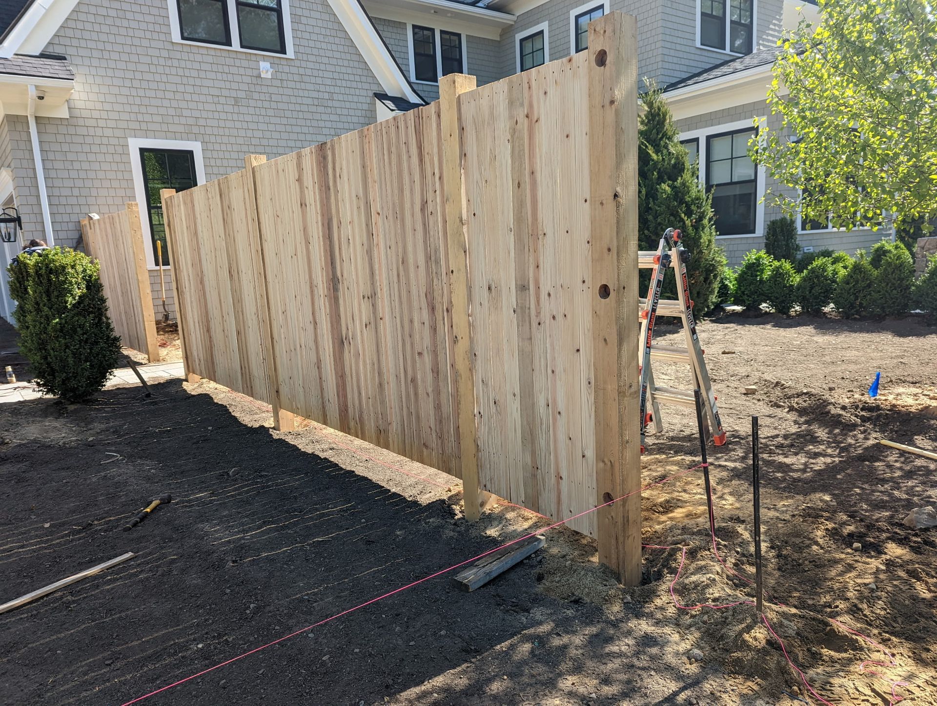A new, light-colored wooden privacy fence stands in a backyard under construction, next to a house with gray siding.