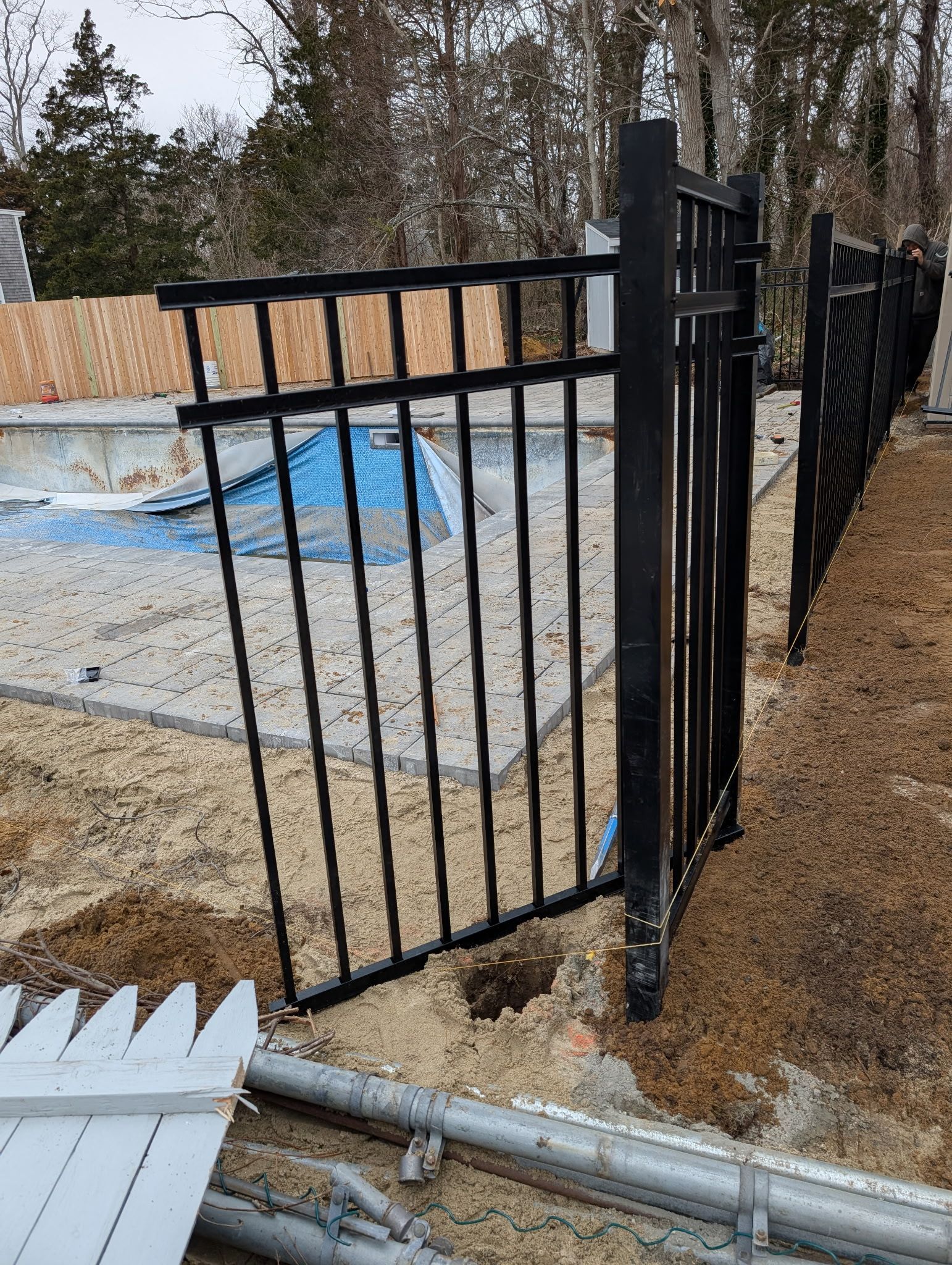 A black metal fence section stands partially installed next to a swimming pool under construction in a wooded yard.