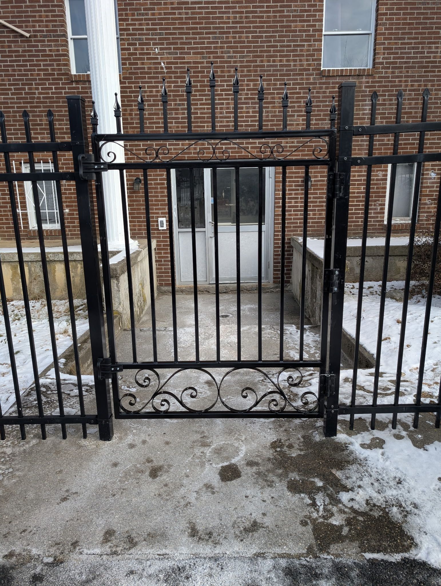 A black wrought iron gate stands in front of a brick building entrance covered in a light dusting of snow.