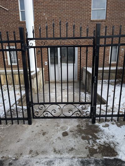 A black metal gate stands before a doorway on a snowy concrete walkway, framed by a brick building.