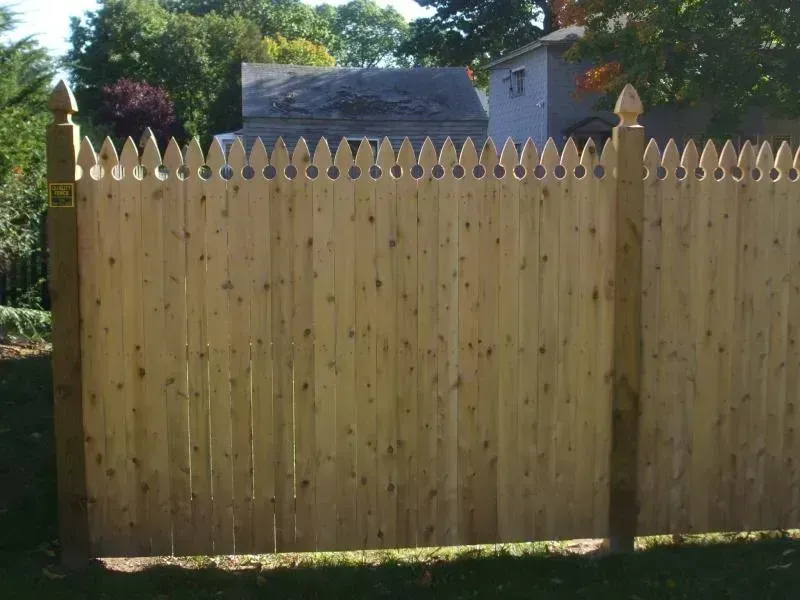 A wooden picket fence with pointed tops and square posts, set against a background of trees and a house.